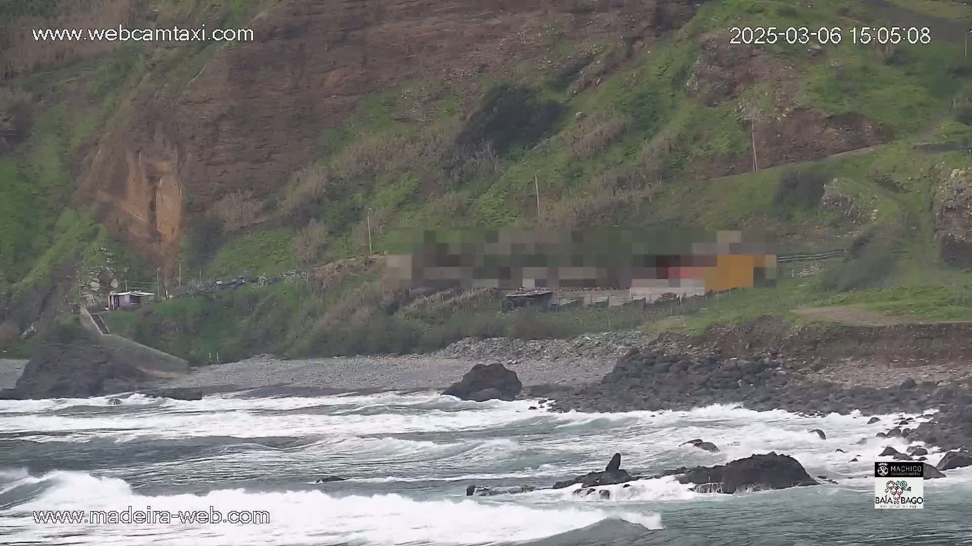 Rough waves crash against a rocky shore next to a green hillside with a few small buildings.