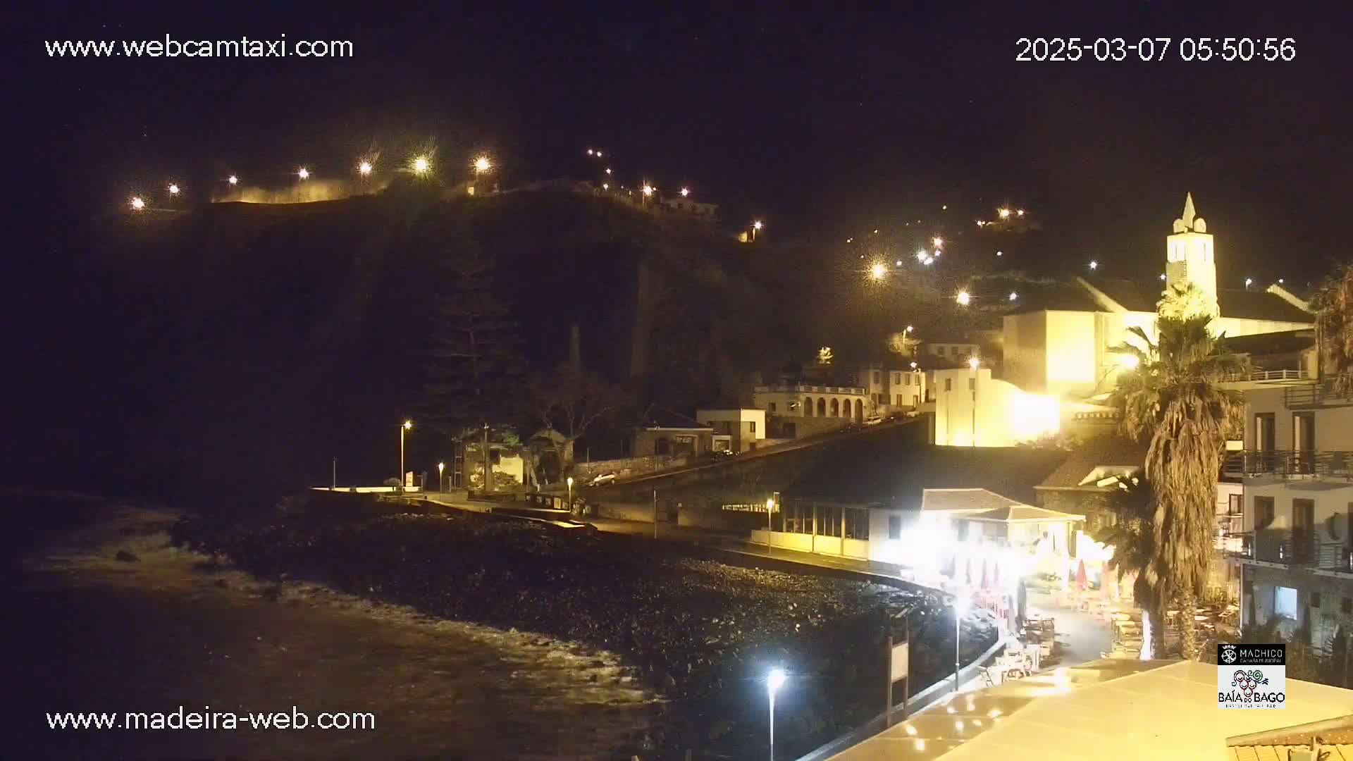 A nighttime coastal town scene with a church steeple, illuminated buildings, and gentle waves lapping the shore.