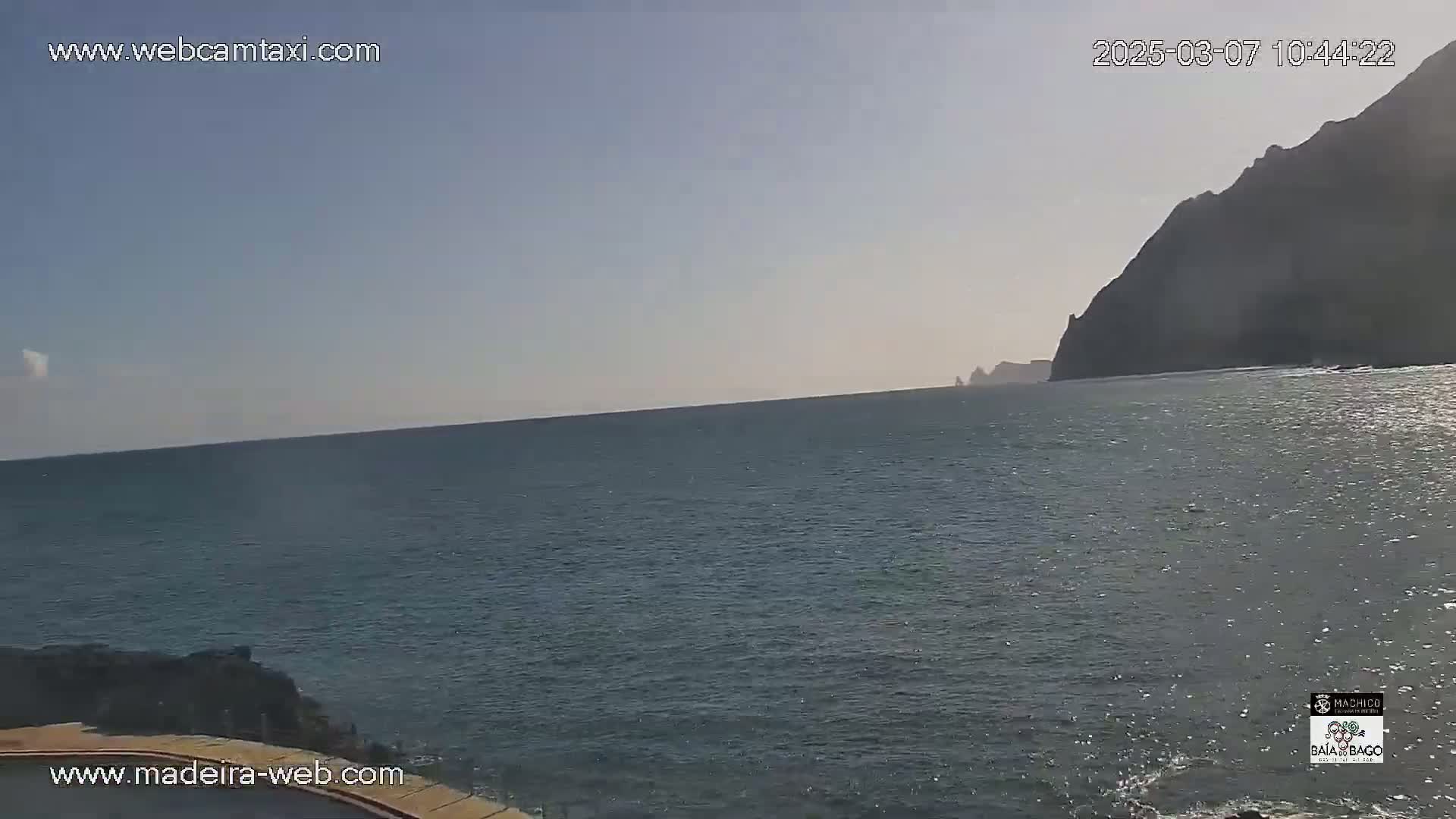 A calm ocean stretches to a dark, rocky cliff face under a clear, bright sky.