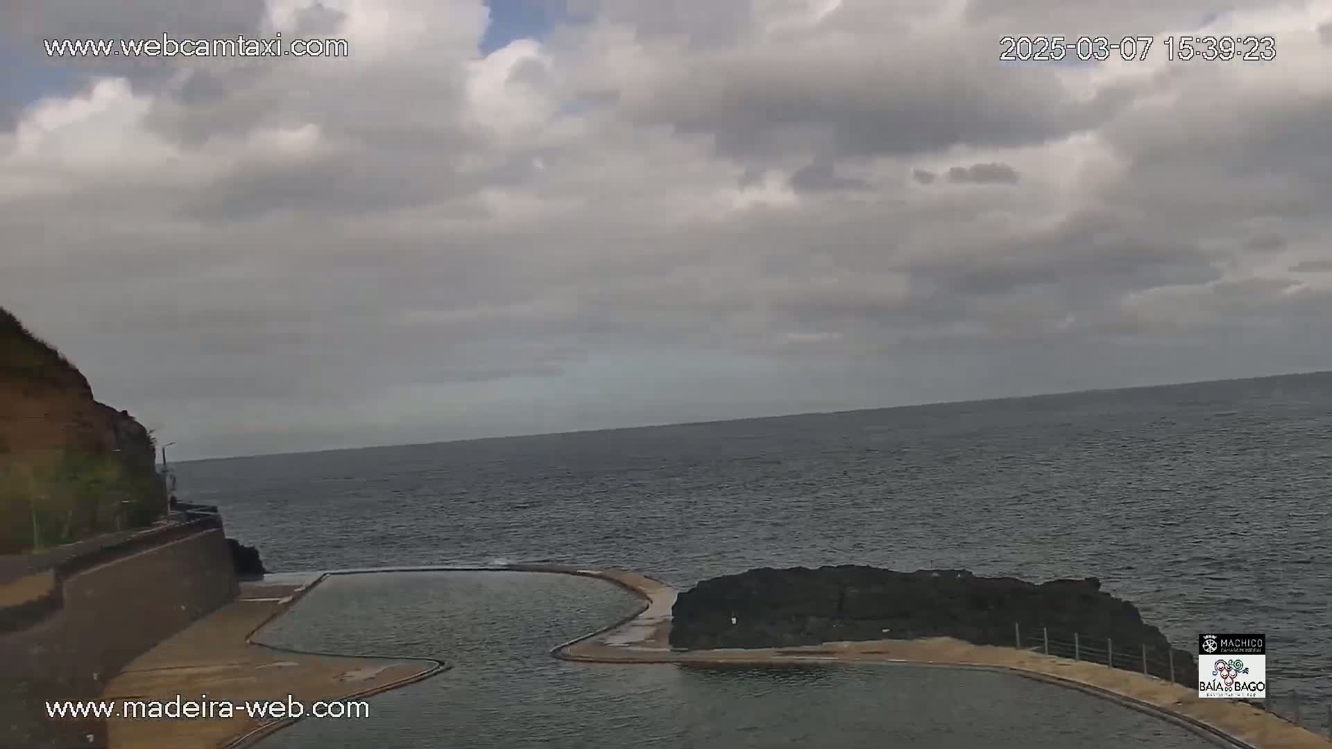 An ocean view shows a partially cloudy sky over a dark body of water with a man-made pool situated on a rocky coast.
