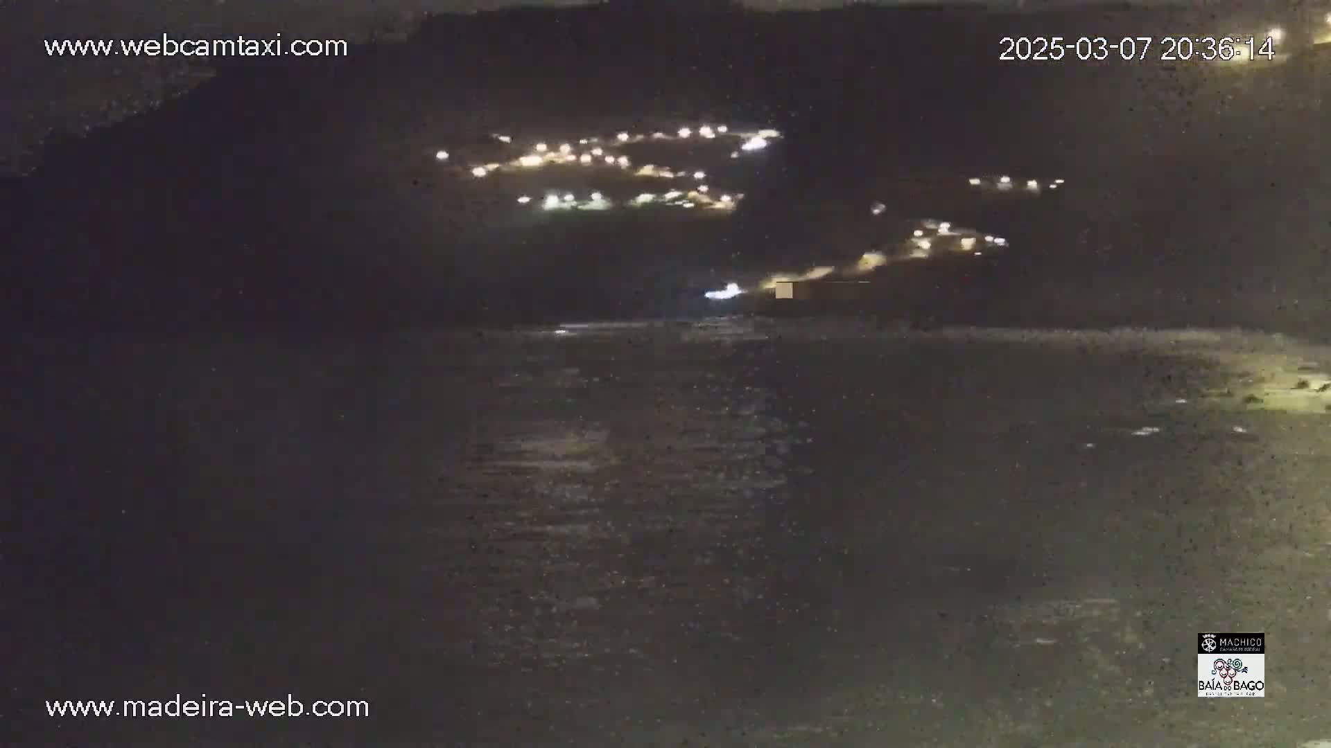 A nighttime, dark view of a body of water with a hillside showing scattered lights in the background.