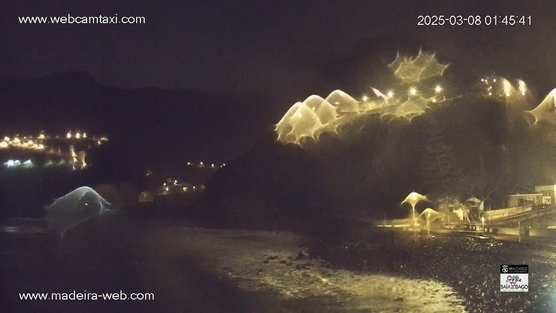 At night, illuminated waves crash on a rocky shore near a hillside with lights visible on structures.
