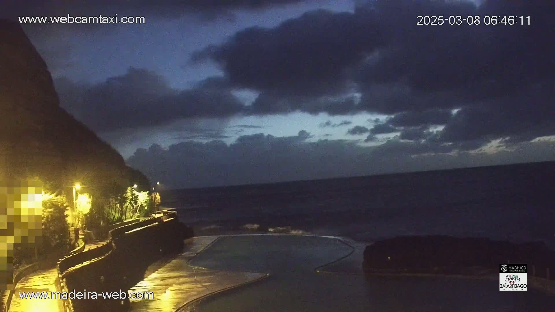 A dark, cloudy night scene shows a coastal area with a dark body of water, a pool, and a paved walkway illuminated by streetlights.