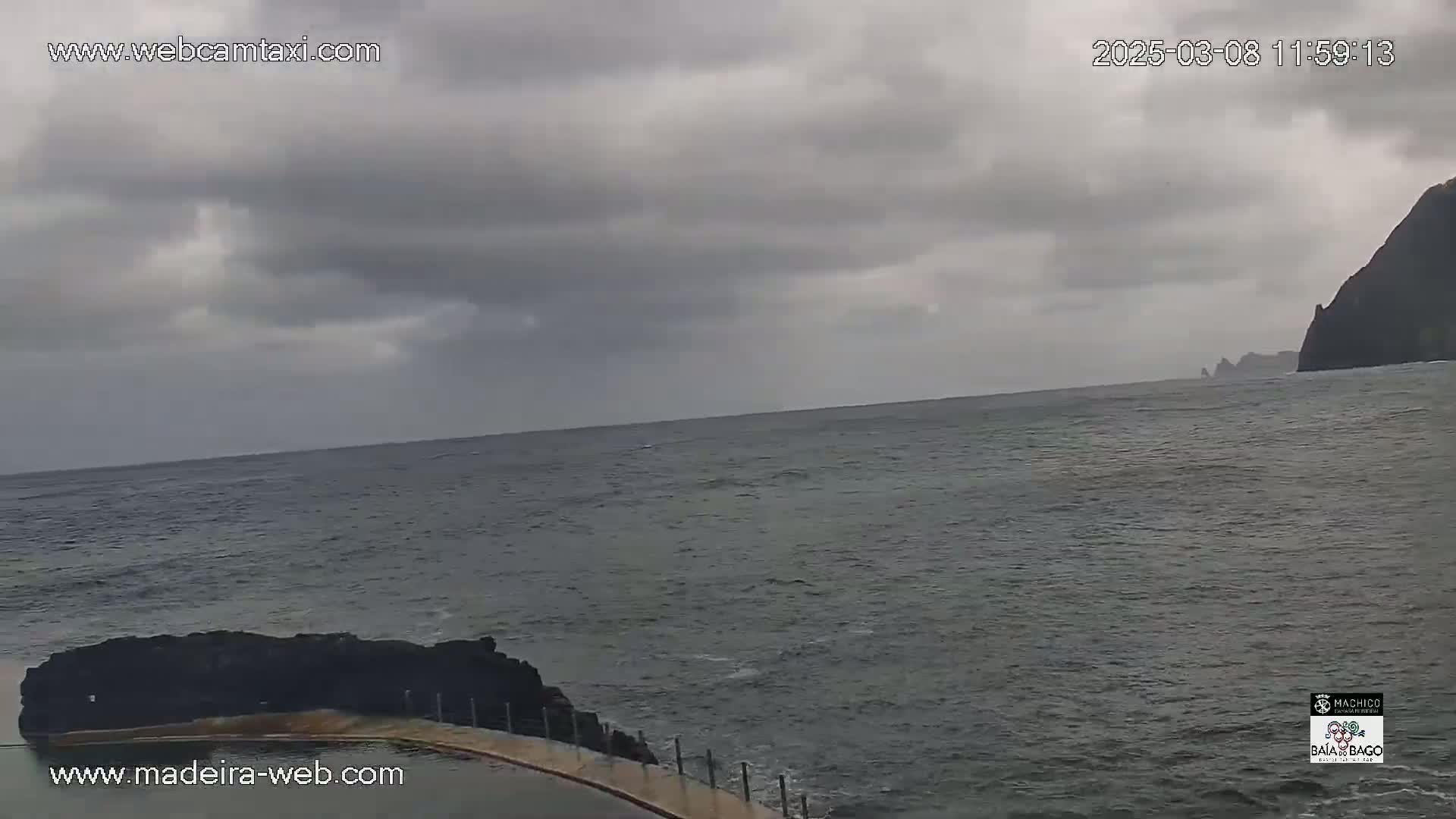 A dark, overcast sky hangs over a gray ocean meeting a rocky shoreline with a partially visible pool, under cloudy skies.