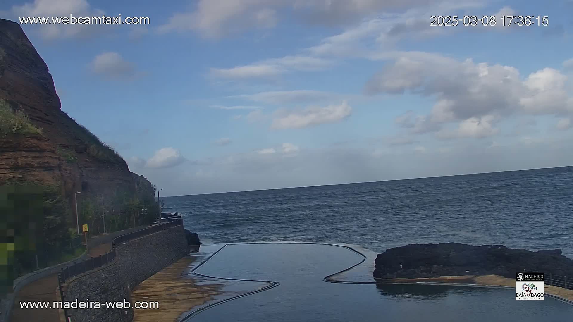 A partly cloudy sky overlooks a rocky coastline with a dark-colored ocean and a dark rectangular pool next to the ocean.