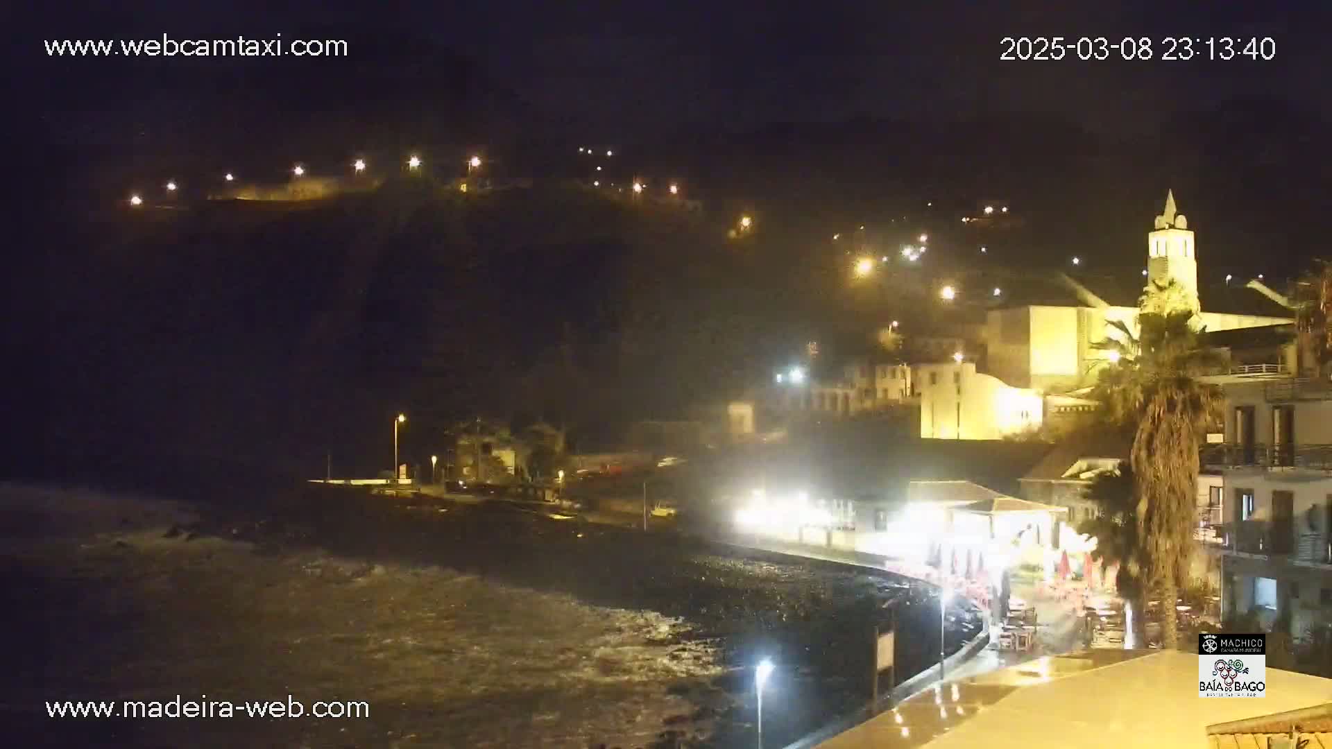 A nighttime coastal town with a church and palm trees is illuminated, with waves crashing on the shore under rainy conditions.