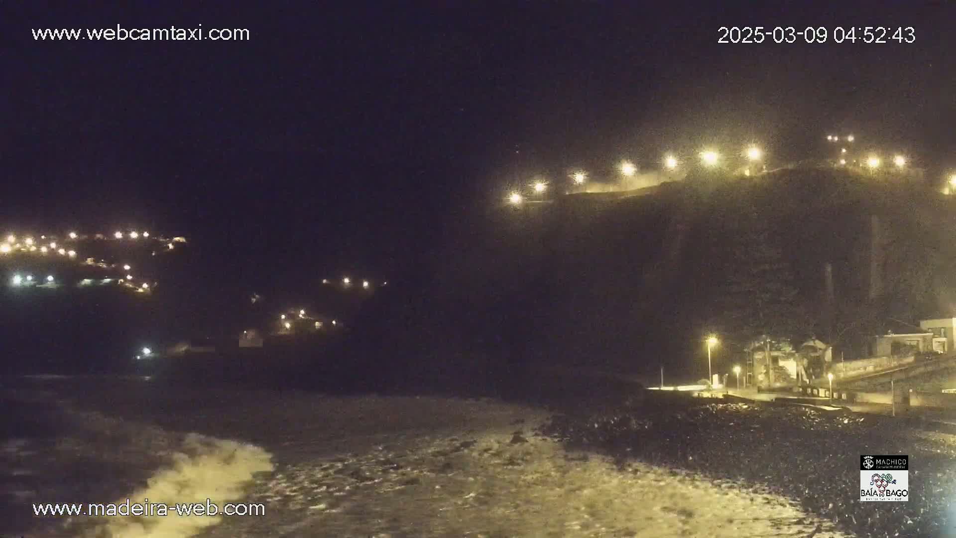 A nighttime coastal scene shows dark, rocky cliffs illuminated by lights, with crashing waves at the base under a clear, dark sky.