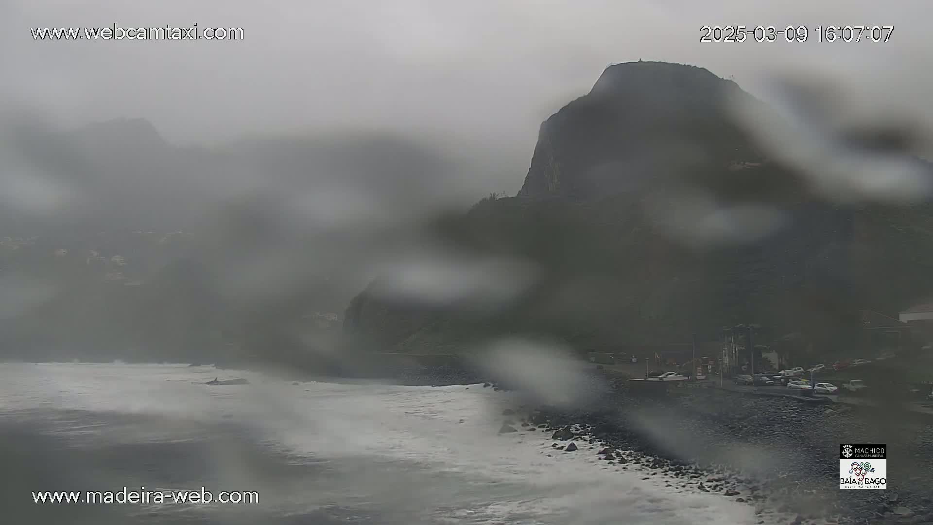 A rain-streaked view shows a rocky coast with waves crashing on the shore near a dark, imposing cliff, under a heavy, overcast sky.
