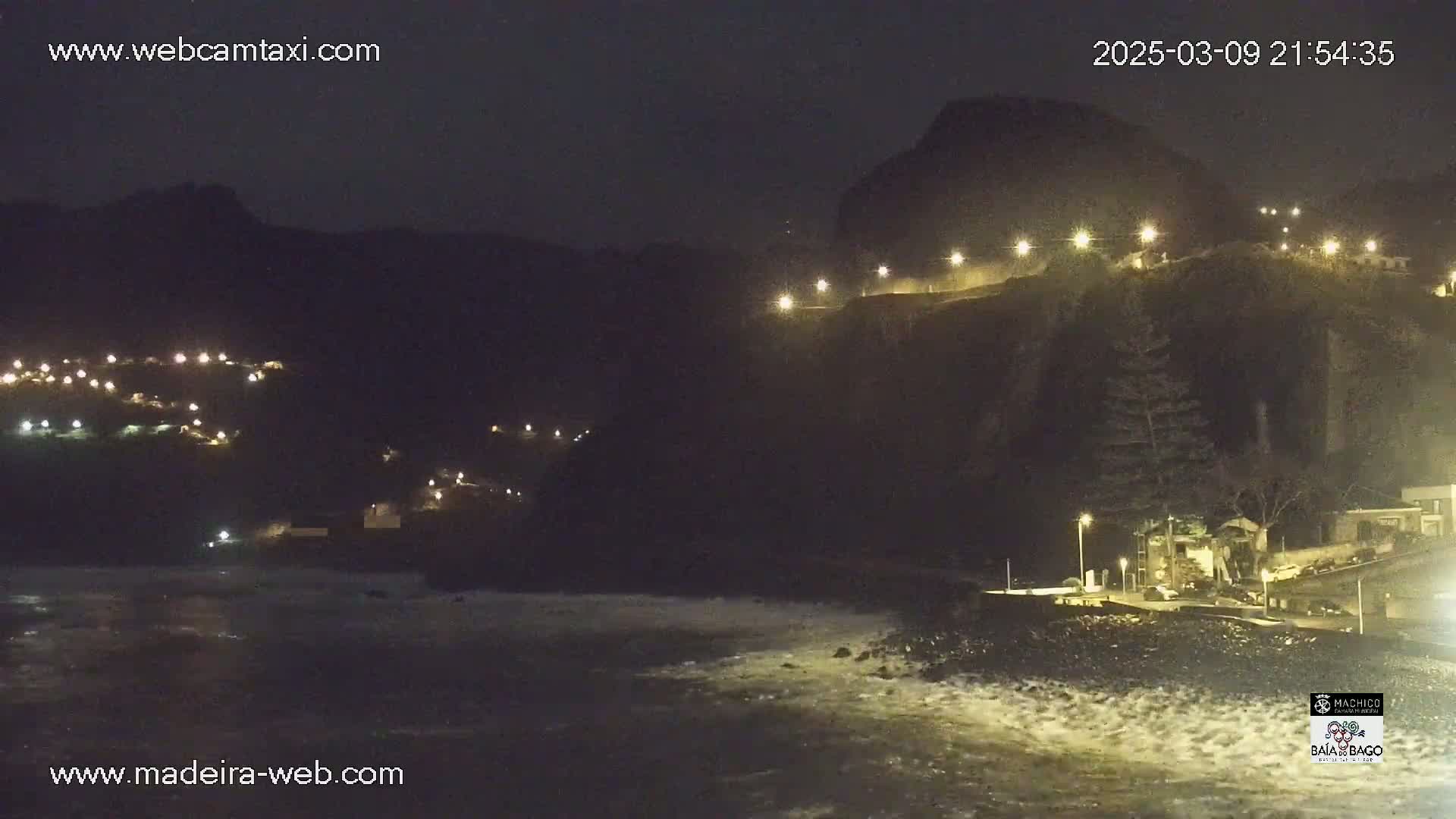 A nighttime coastal scene shows a dark, rocky cliff with lights illuminating a path along its top, a village with scattered lights below, and a dark ocean with small waves crashing on the shore.
