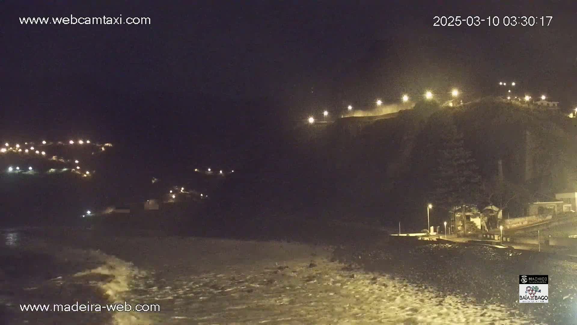 A nighttime coastal scene shows a dark cliff illuminated by lights, with waves breaking on the shore below.