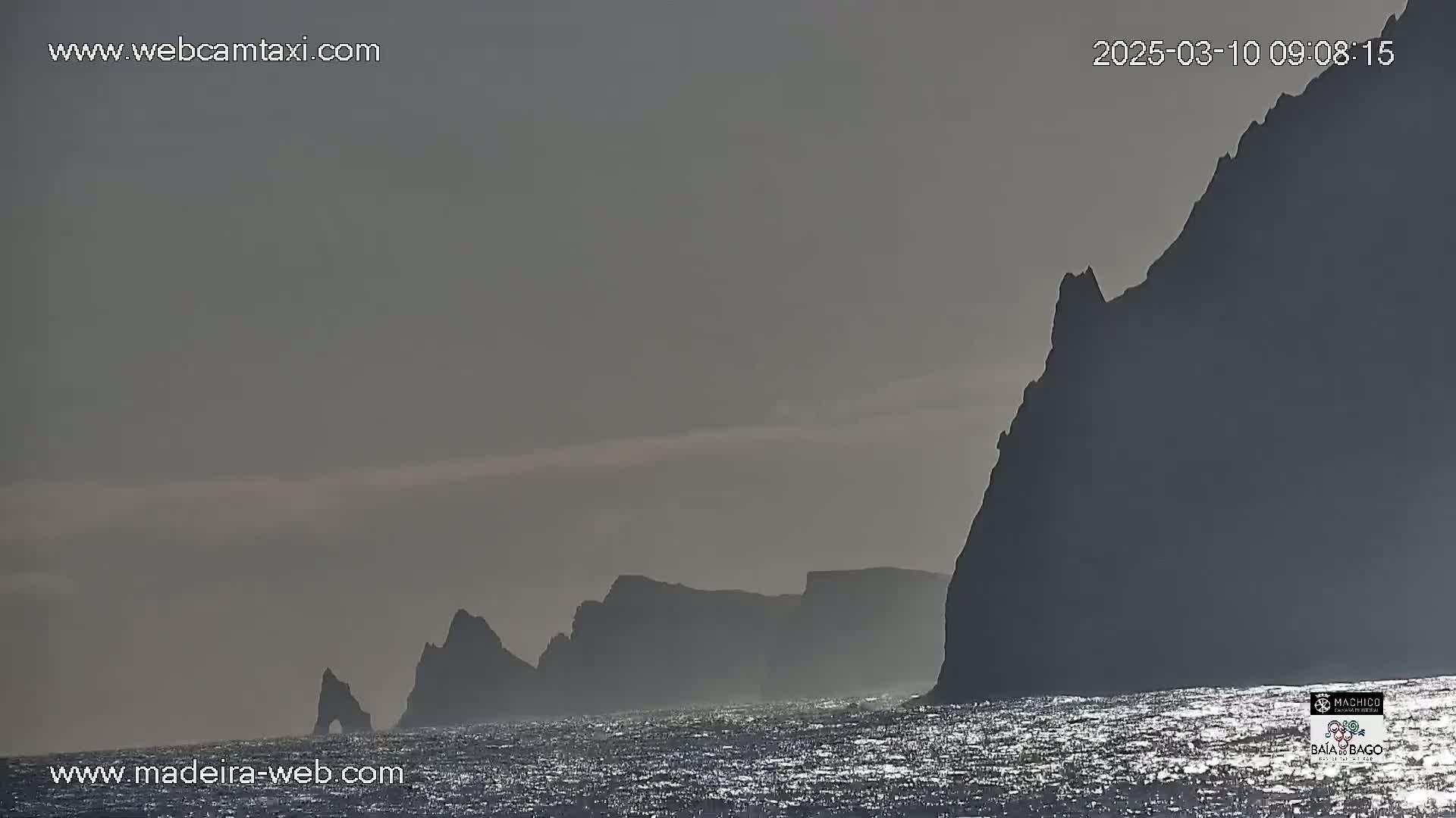 Silhouetted cliffs rise from a calm, glittering ocean under a hazy sky.