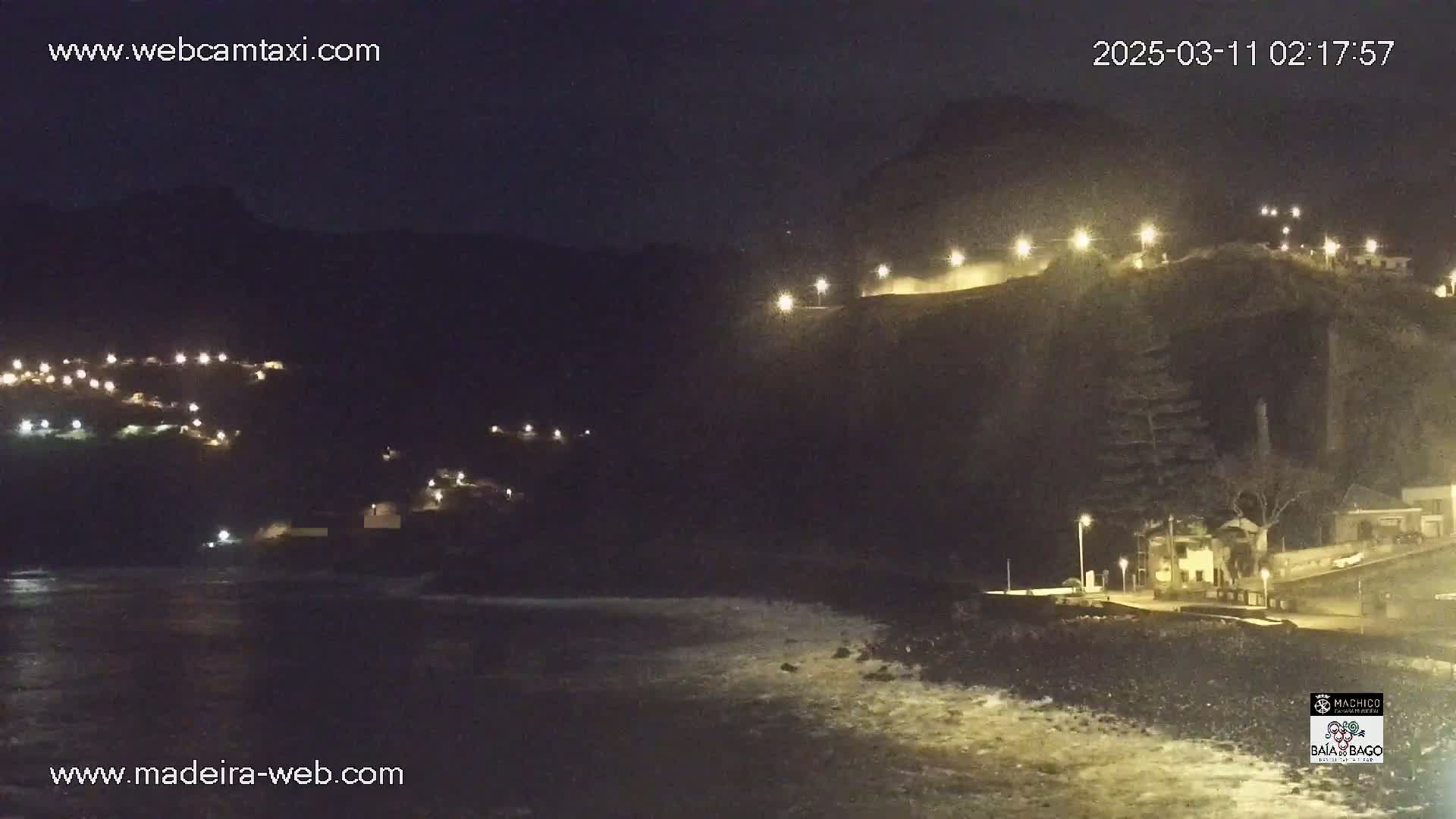 A nighttime coastal scene shows a dark cliff face with lights illuminating a road and buildings, while waves crash on the shore below.