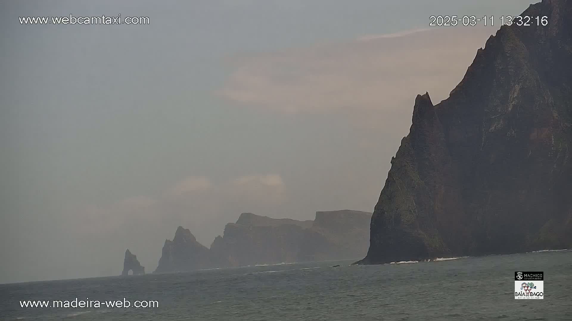 Dark, imposing cliffs rise from a gray, somewhat hazy ocean under an overcast sky.