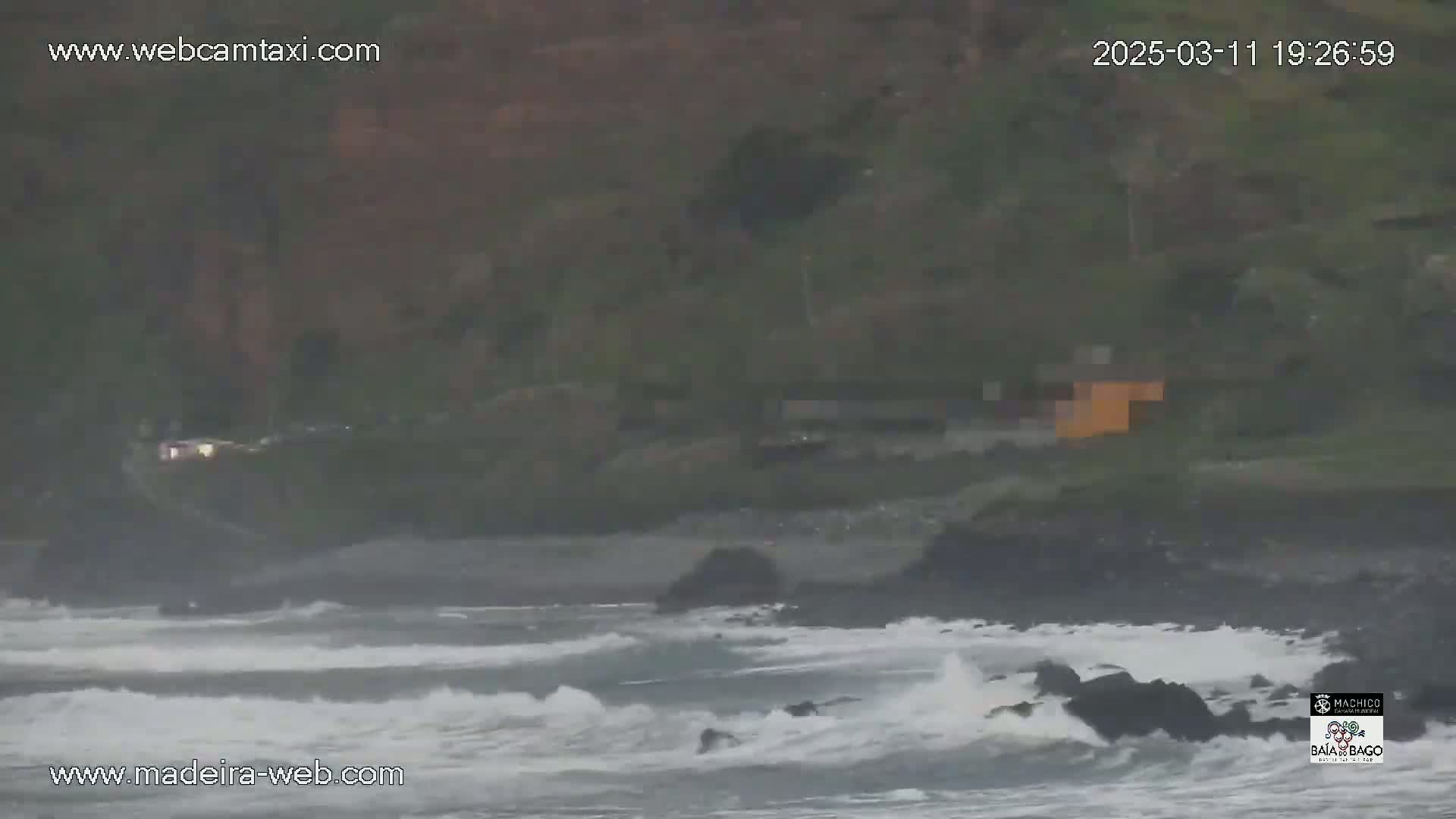 Dark, overcast conditions show rough waves crashing against a rocky shoreline near a cliffside with some structures visible.