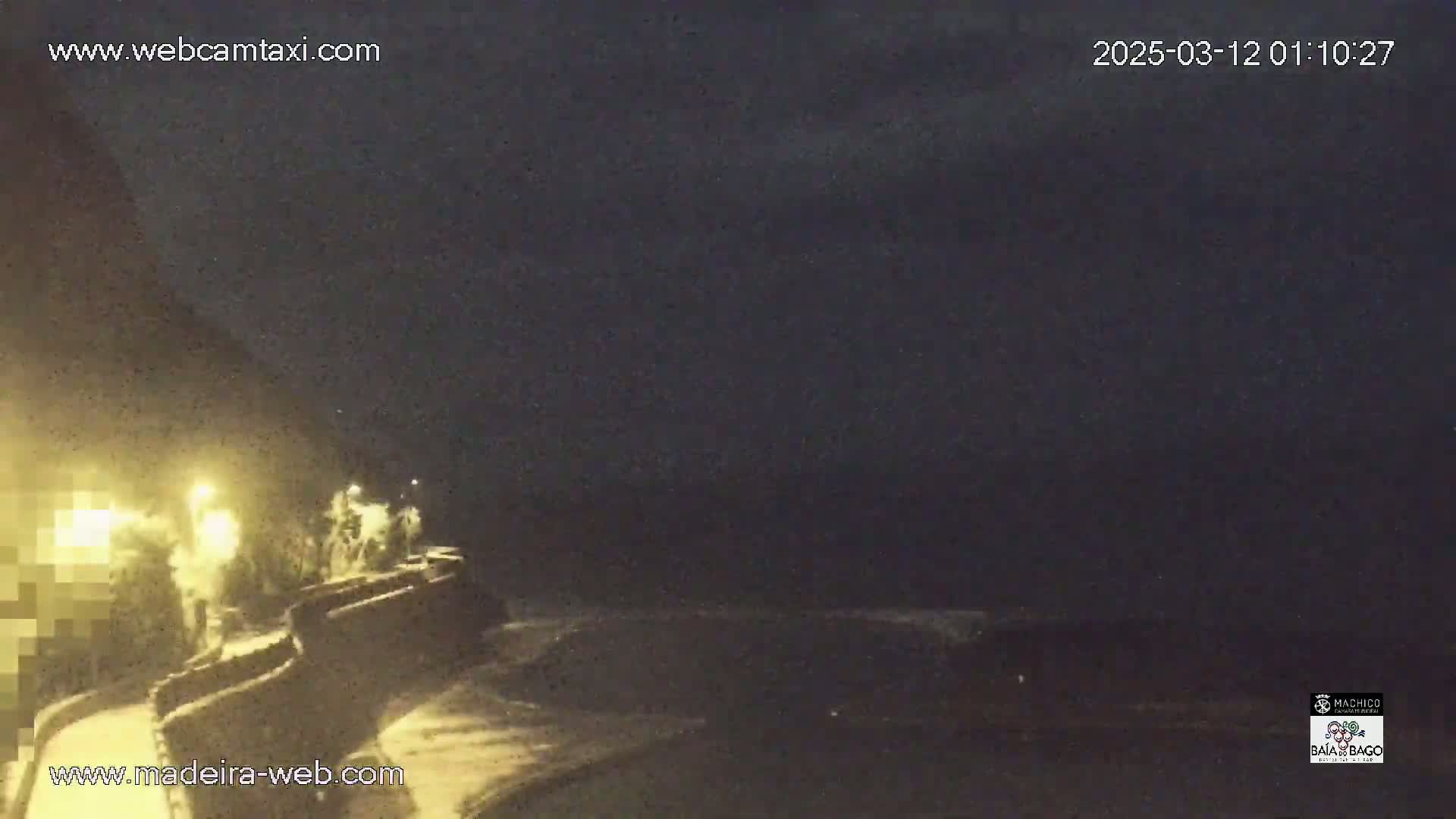 A nighttime, low-light view of a coastal area shows a dark ocean, a dimly lit roadway, and several figures near a wall.