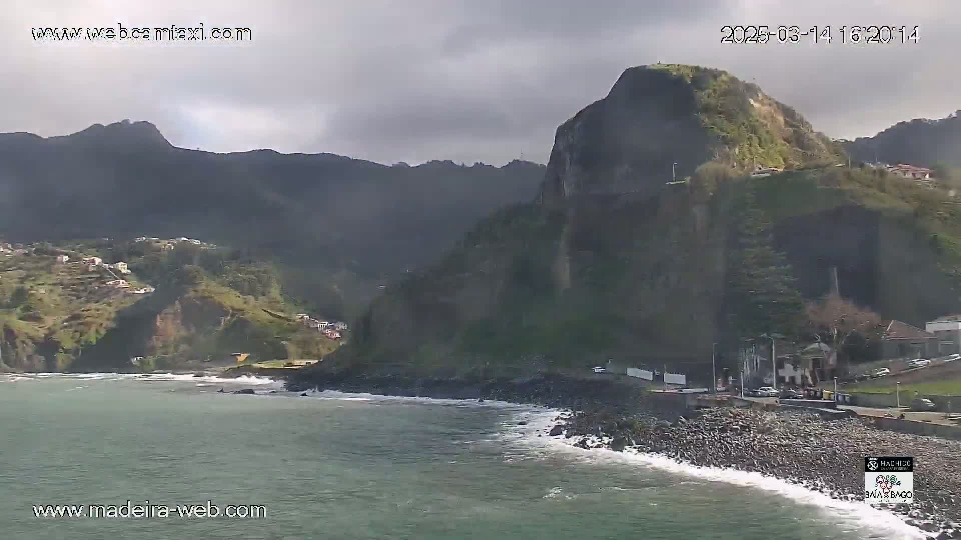 A rocky coastline with dark green mountains in the background under a cloudy sky, with a small town visible along the shore.