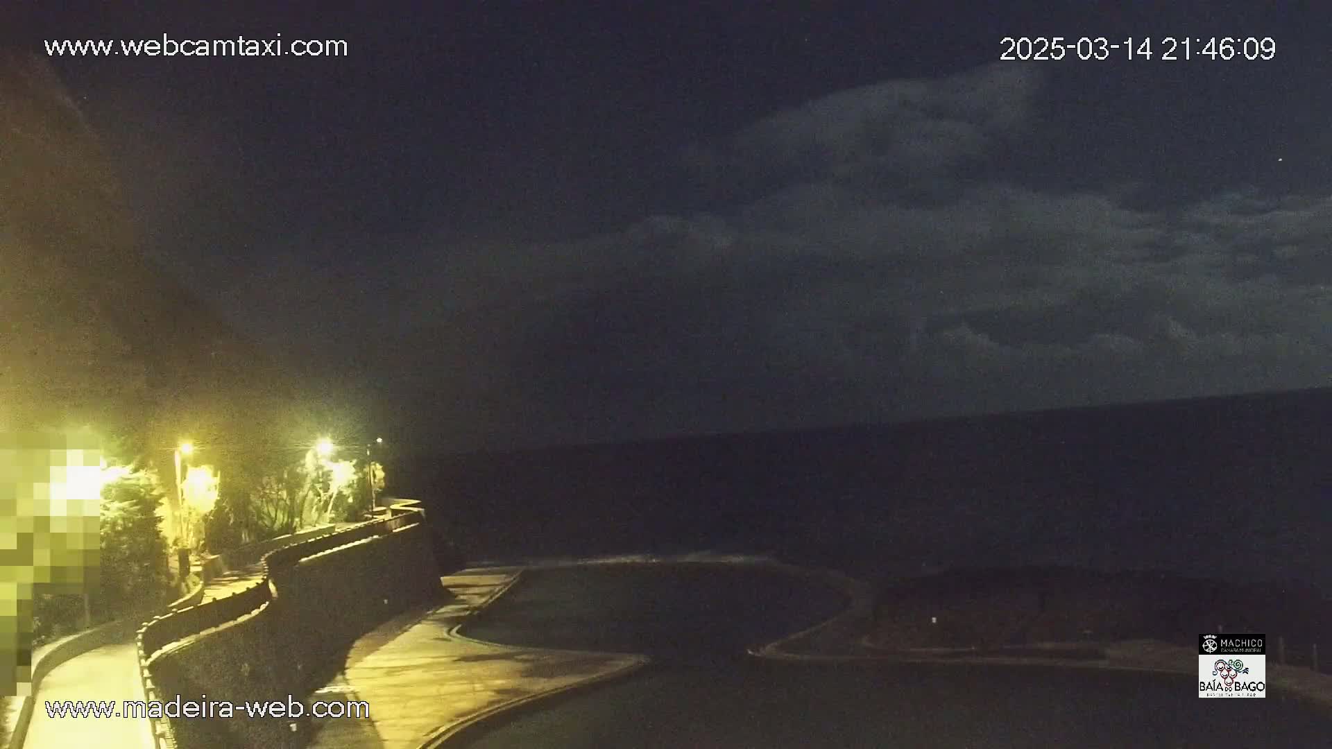 A nighttime view of a dark ocean under a partly cloudy sky, with a curved walkway and pool lit by several lamps along a cliffside.