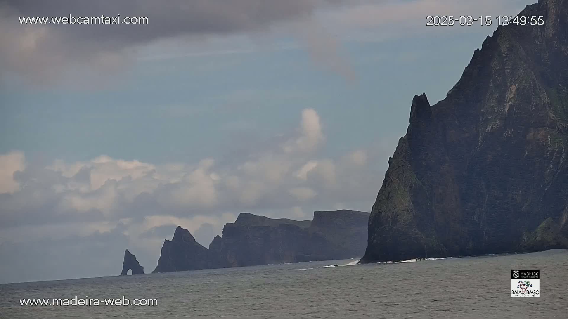 Dark, rocky cliffs rise from a calm ocean under a mostly cloudy sky.
