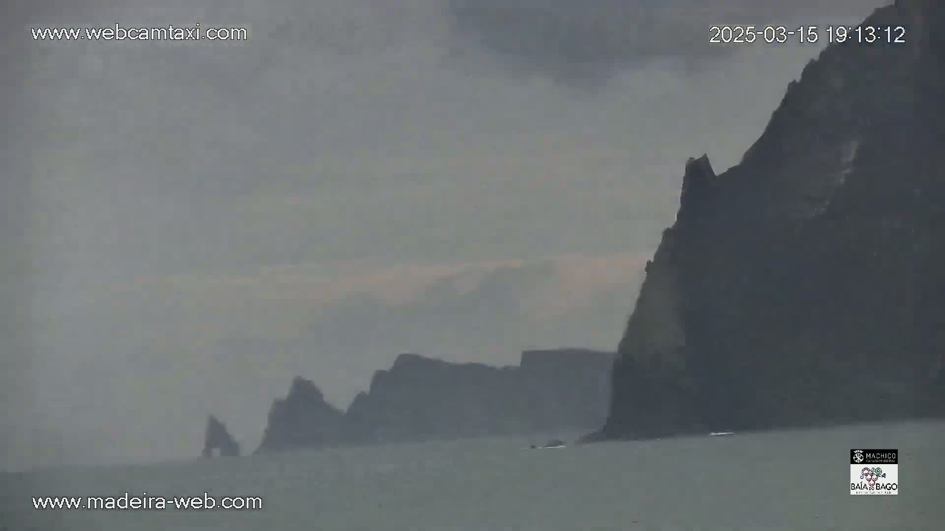 Dark, rocky cliffs rise from a calm, grey sea under an overcast sky.