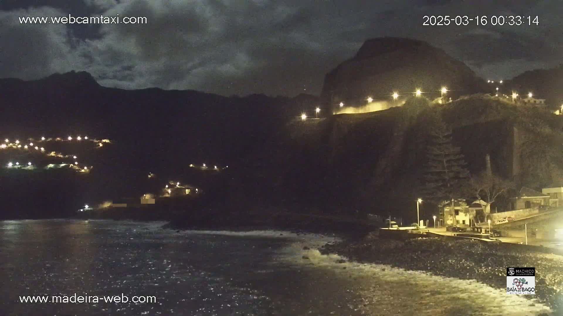 A nighttime coastal scene shows dark, cloudy skies over a rocky shoreline with sparsely lit buildings and a dark mountain in the background, and slightly wavy water in the foreground.