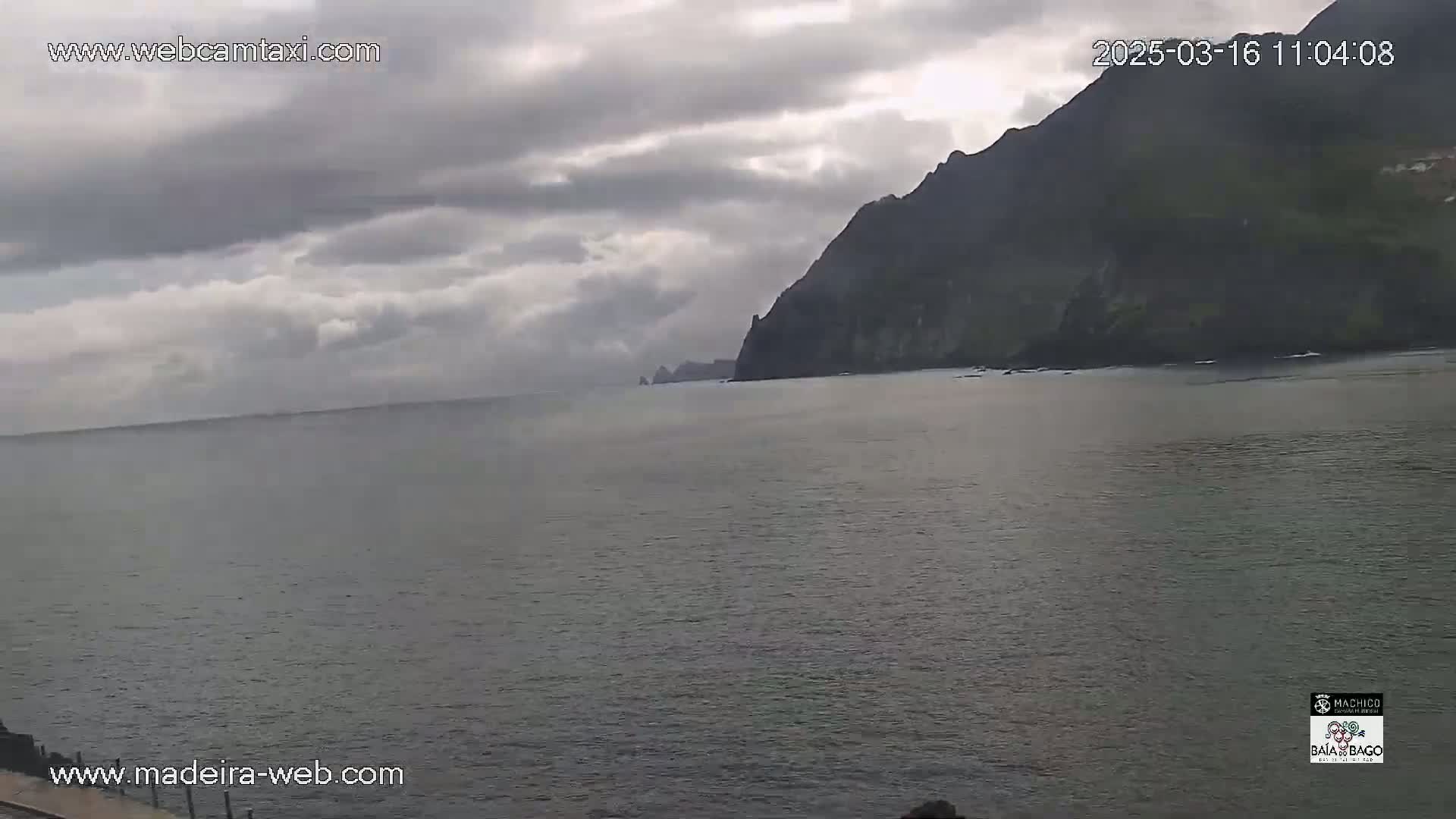 A calm, grey ocean stretches out to meet a dark, rocky cliff under a cloudy sky.