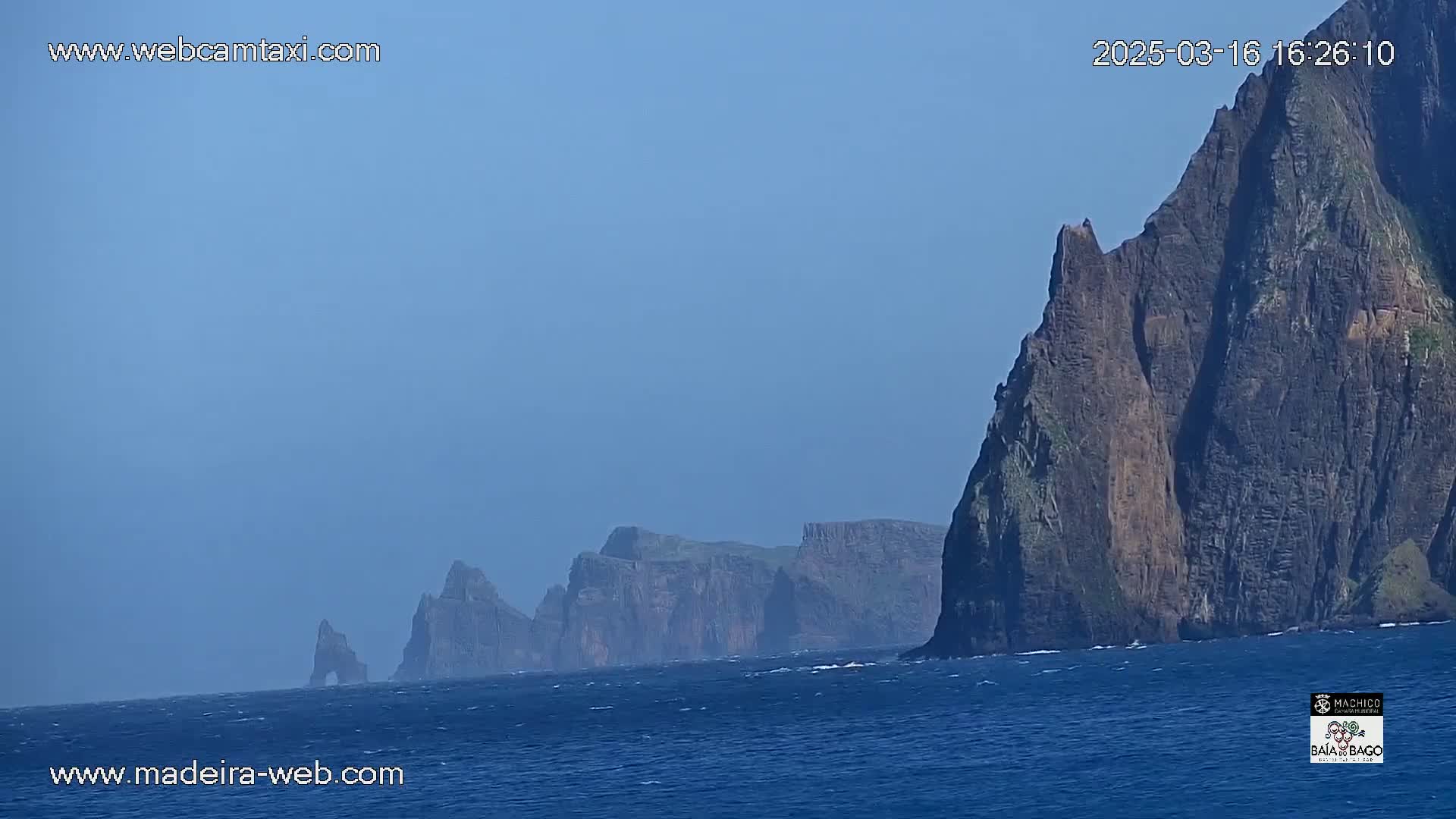 A hazy, blue sky overlooks dark, rocky cliffs rising from a choppy ocean.