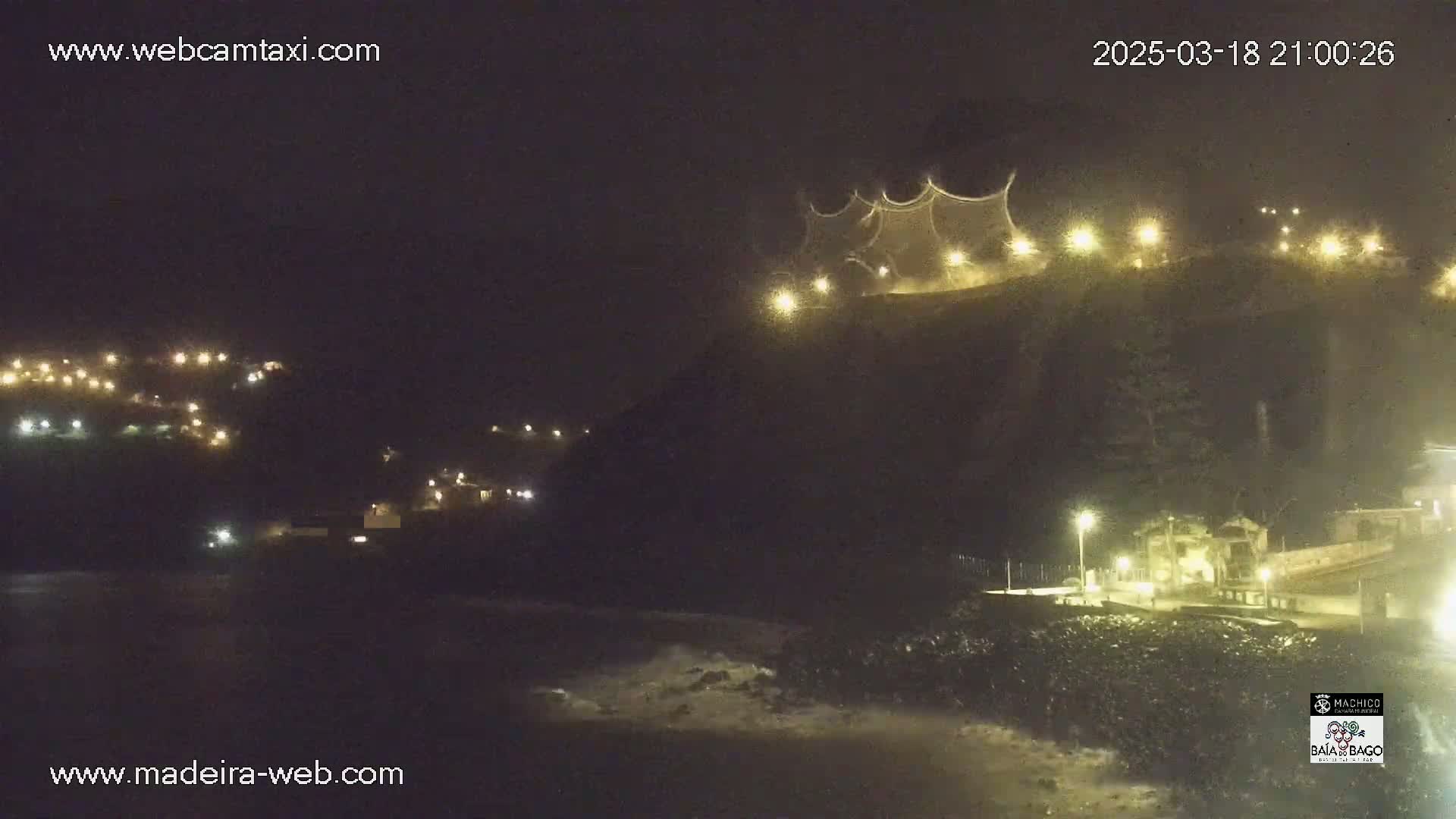 A nighttime coastal scene shows a dark cliff face with a lighted structure at its peak, and houses with lights illuminating the lower slopes, with dark ocean waves gently lapping the shore below.