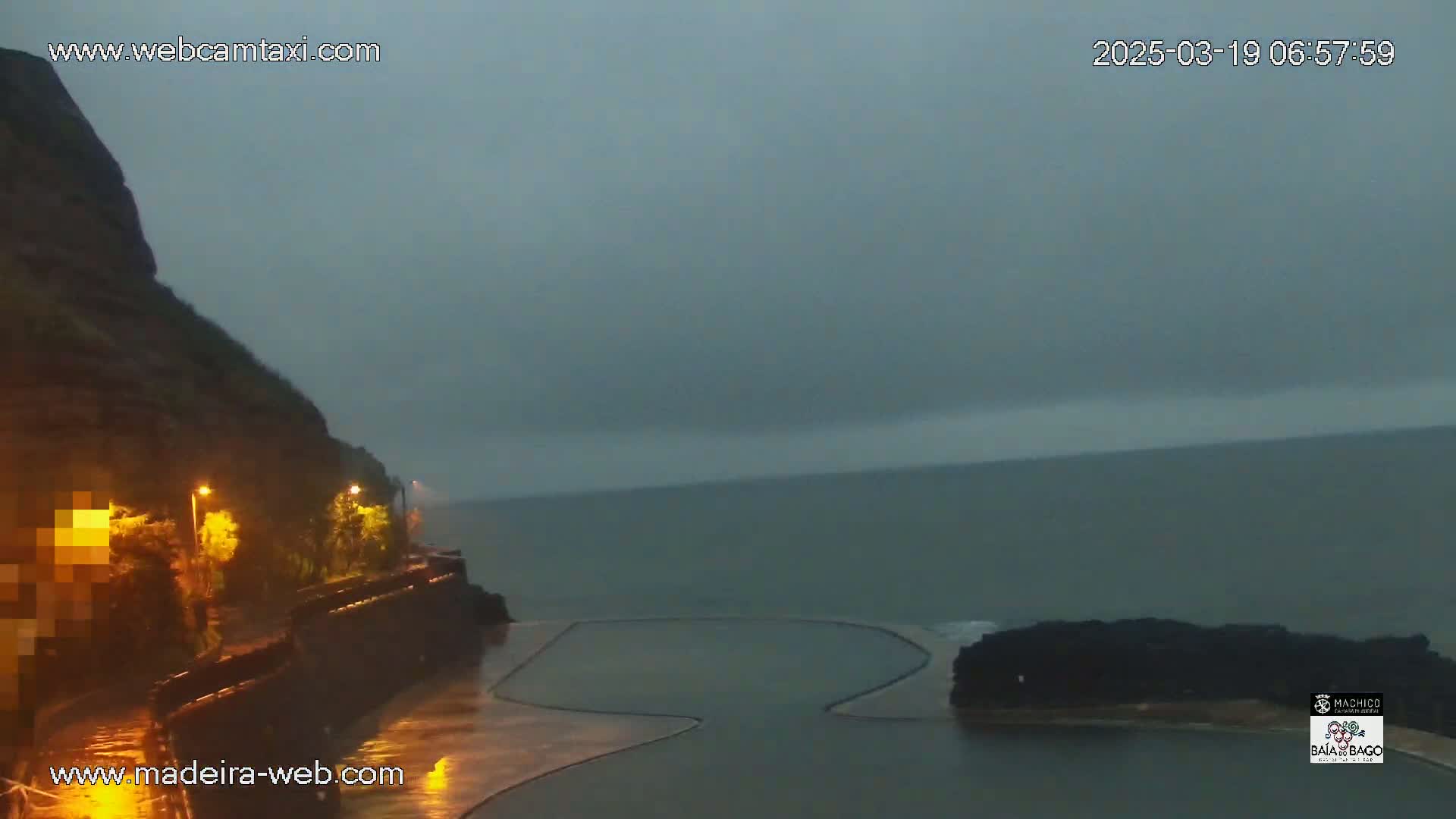 A dark, overcast sky hangs over a wet coastal scene featuring a rocky cliff, lit streetlights, and a partially visible, empty ocean swimming pool.
