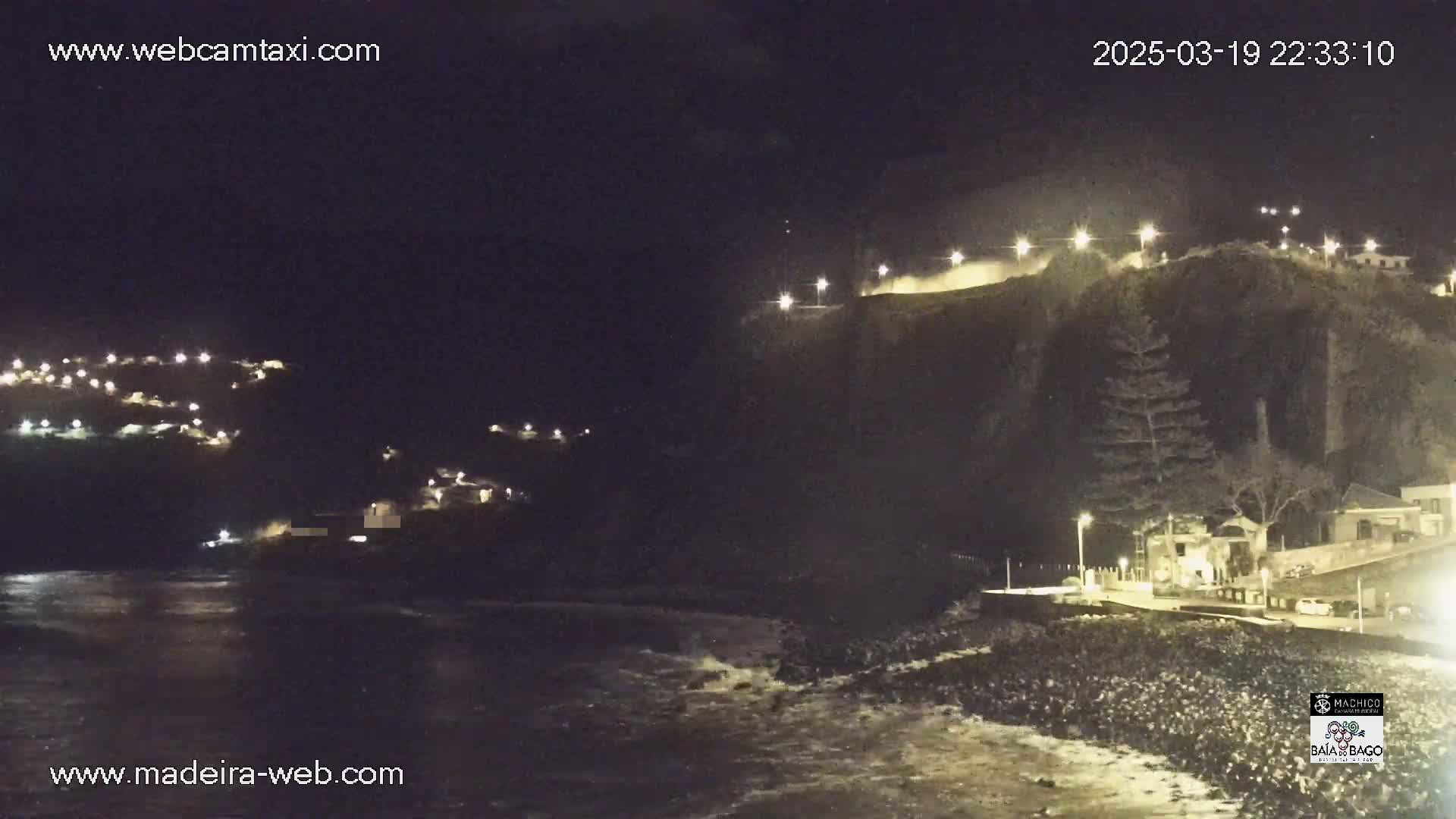 A nighttime coastal scene shows a dark cliff face with lights illuminating a roadway and buildings, while a dark ocean with small waves laps at the shore below.