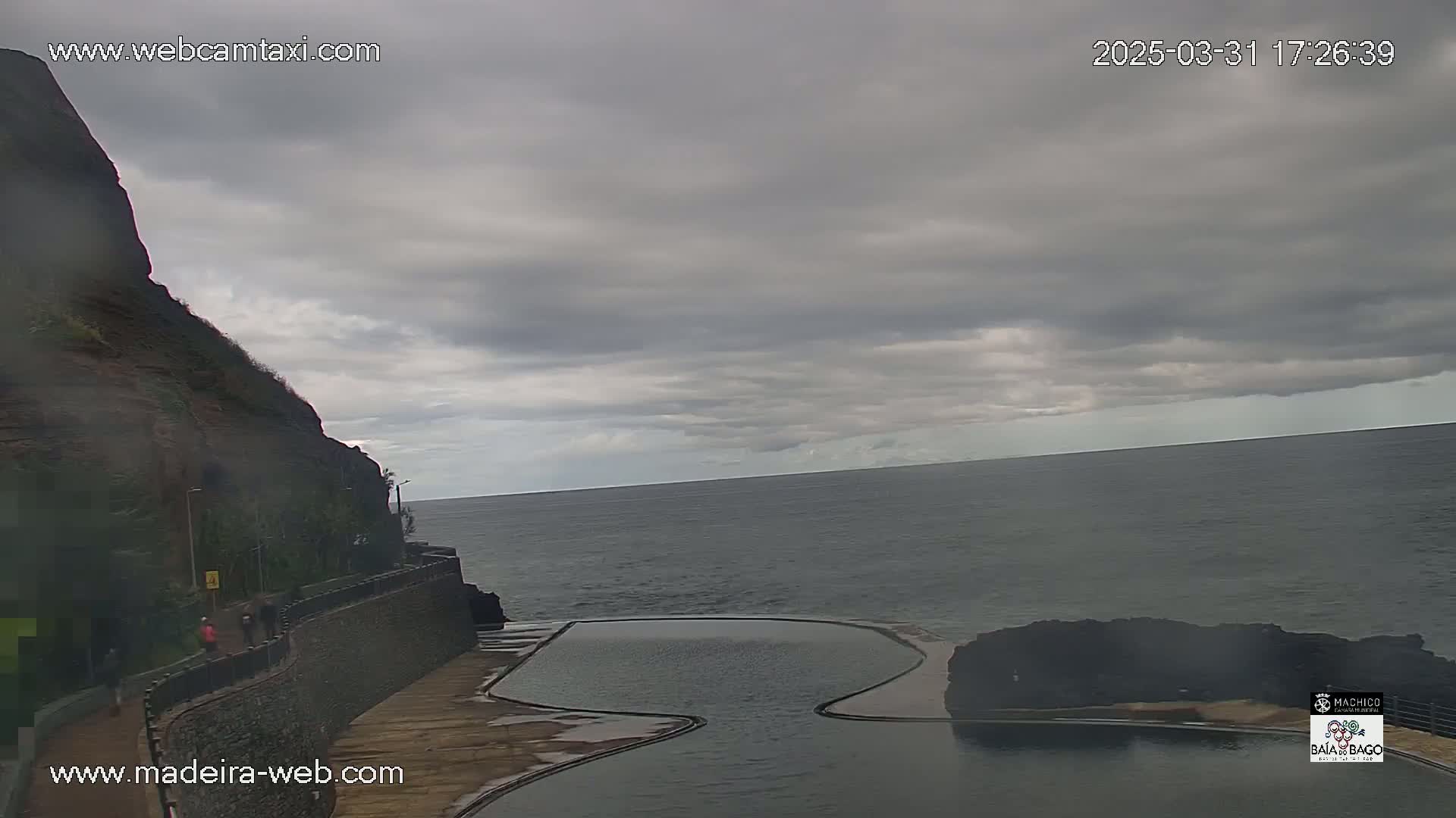 A rocky cliff overlooks a dark ocean with an empty, irregular-shaped pool situated between the cliff and the water under an overcast sky.