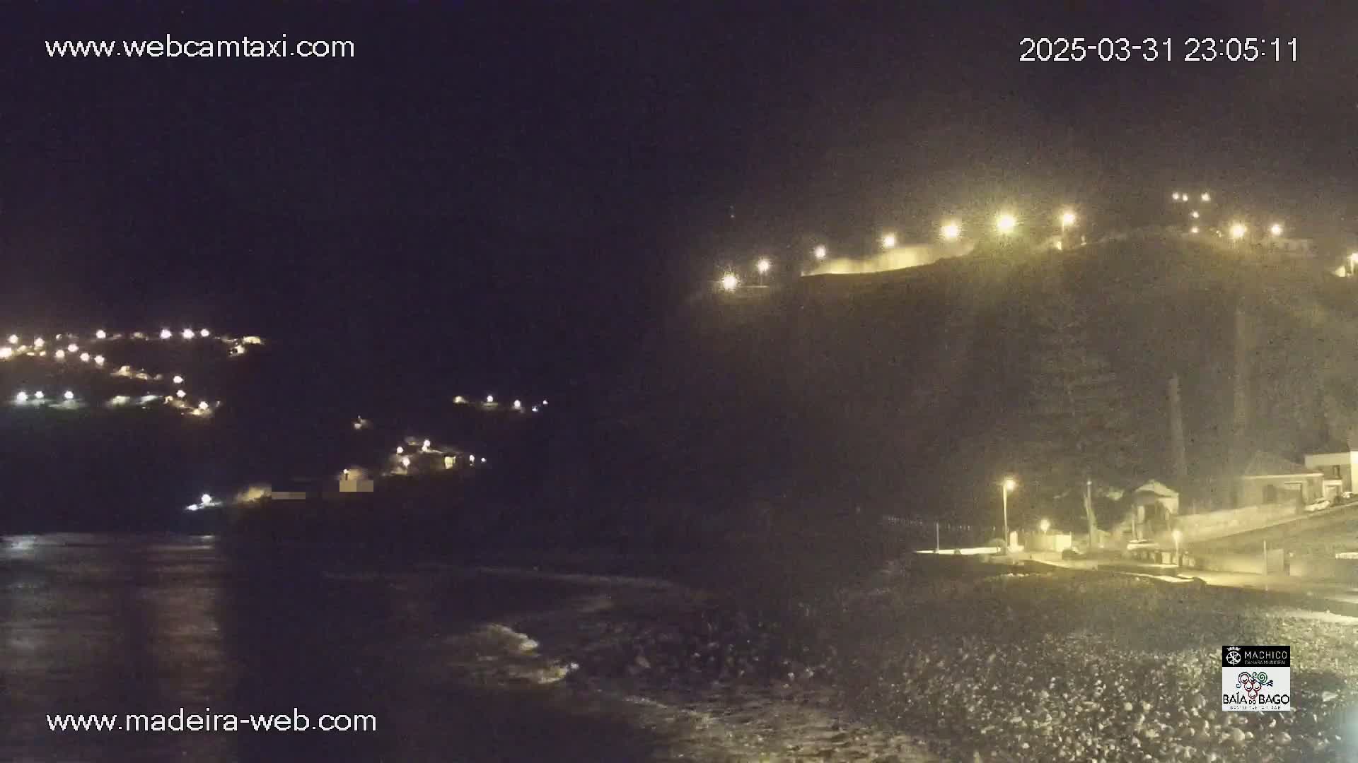 A nighttime coastal scene shows a dark, rocky shoreline with gentle waves, illuminated buildings on hillsides, and lights along a waterfront path.