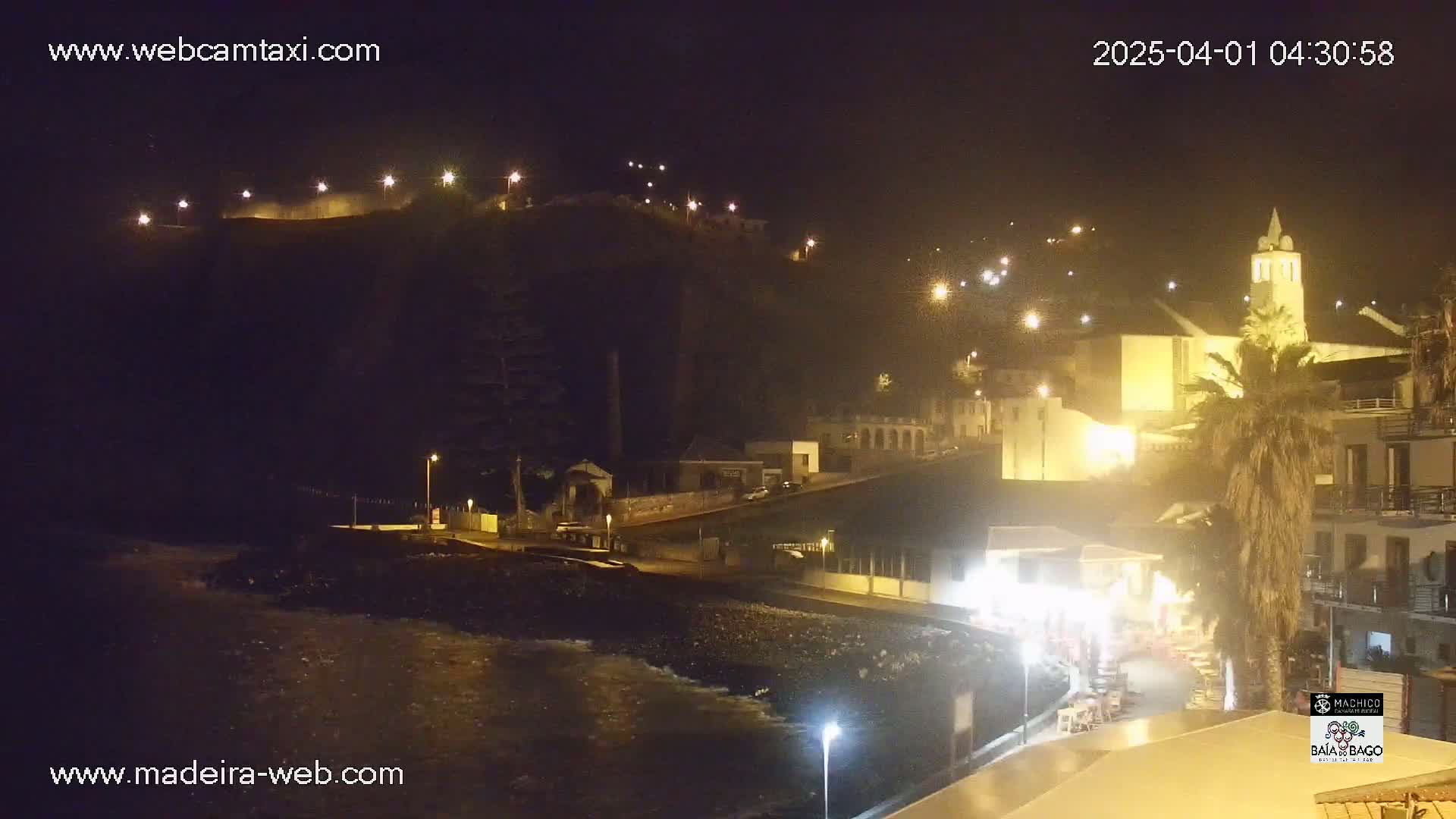 A nighttime coastal town scene shows a dark hilltop illuminated by lights, buildings along a shoreline, and a body of water with gentle waves.