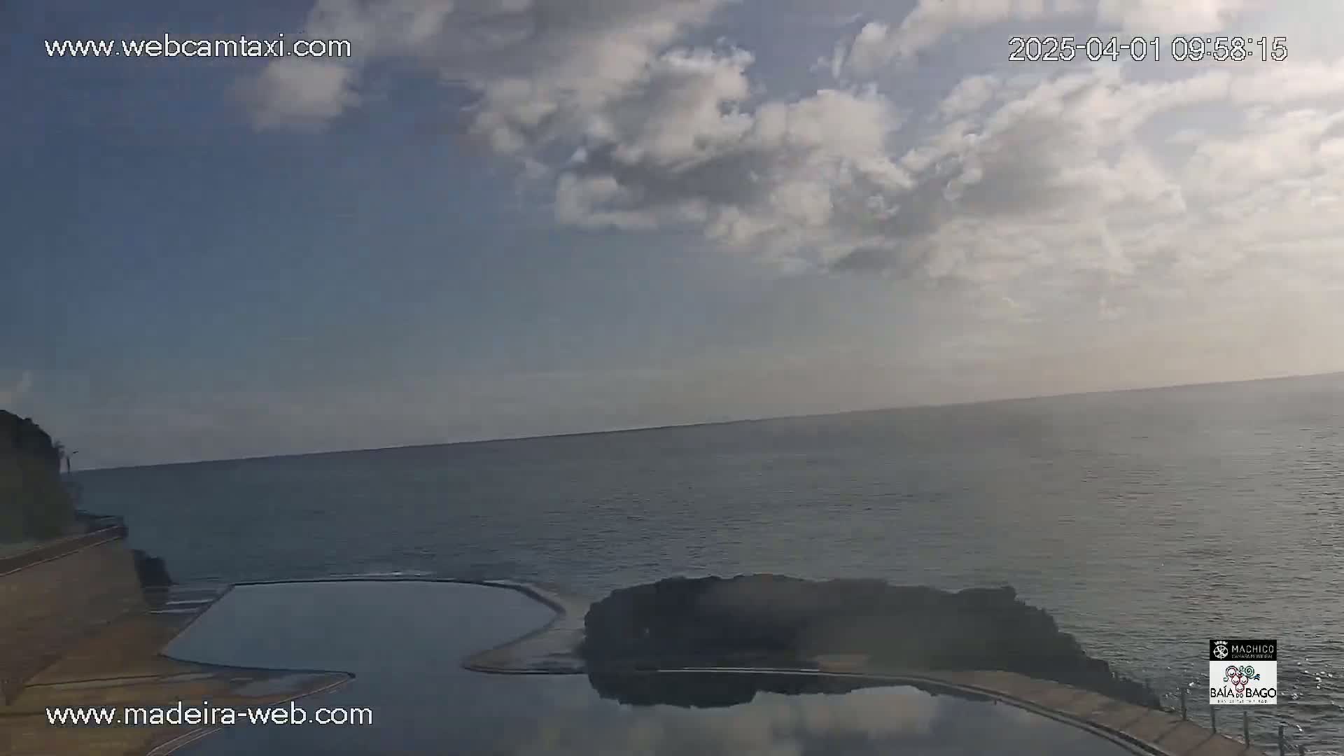 An outdoor view shows a partly cloudy sky over a calm ocean, with a dark-colored infinity pool situated on a rocky cliff overlooking the water.