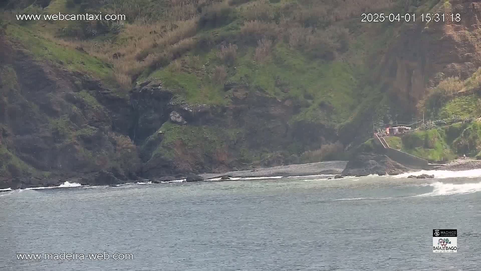 A rocky, green hillside slopes down to a small, grey-sand beach where small waves are breaking onto the shore on a partly sunny day.