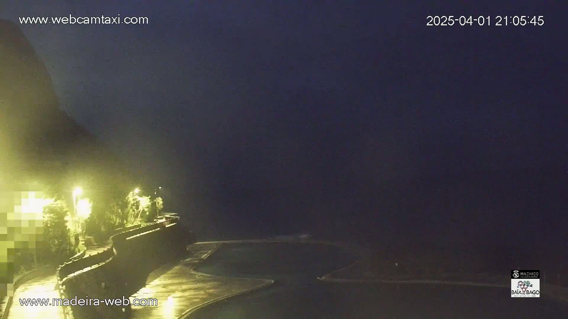 A nighttime view of a coastal area with a dark, possibly foggy or hazy sky, showing a curving roadway lit by lamps and a dark body of water below.