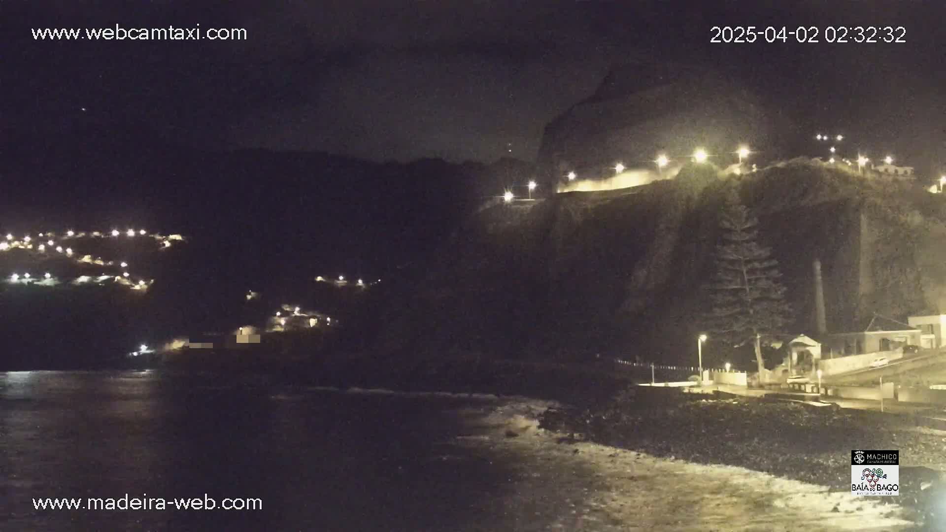 A nighttime coastal scene shows a dark, rocky cliff with lights illuminating a road and buildings, overlooking a dark ocean with small waves.
