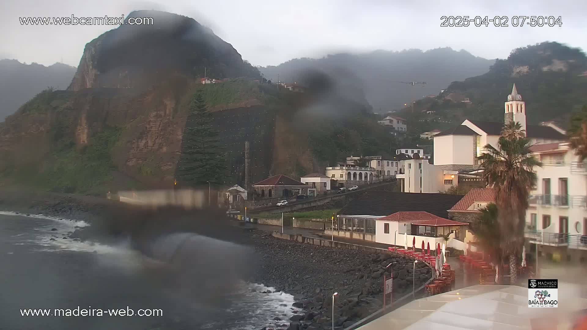 A rain-swept coastal town nestled against a steep, rocky cliffside, with waves crashing on the shore.