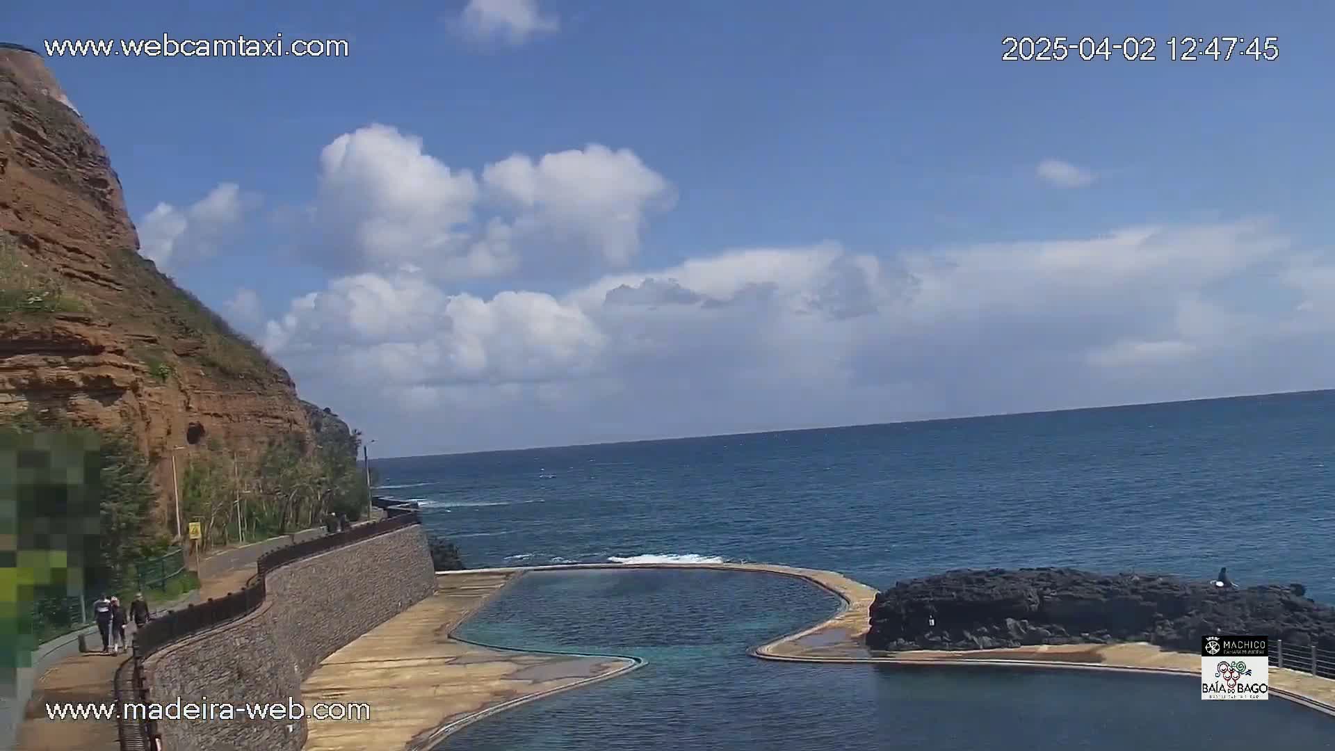 A partly cloudy day shows a rocky cliff overlooking a calm ocean with a natural seawater pool built into the rocks.