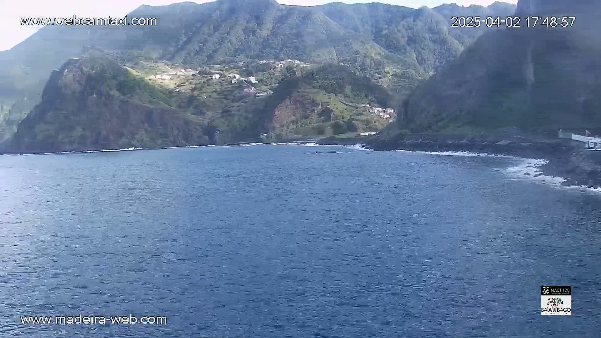 A calm, blue ocean laps against a rocky shore beneath verdant, mountainous terrain dotted with small buildings.