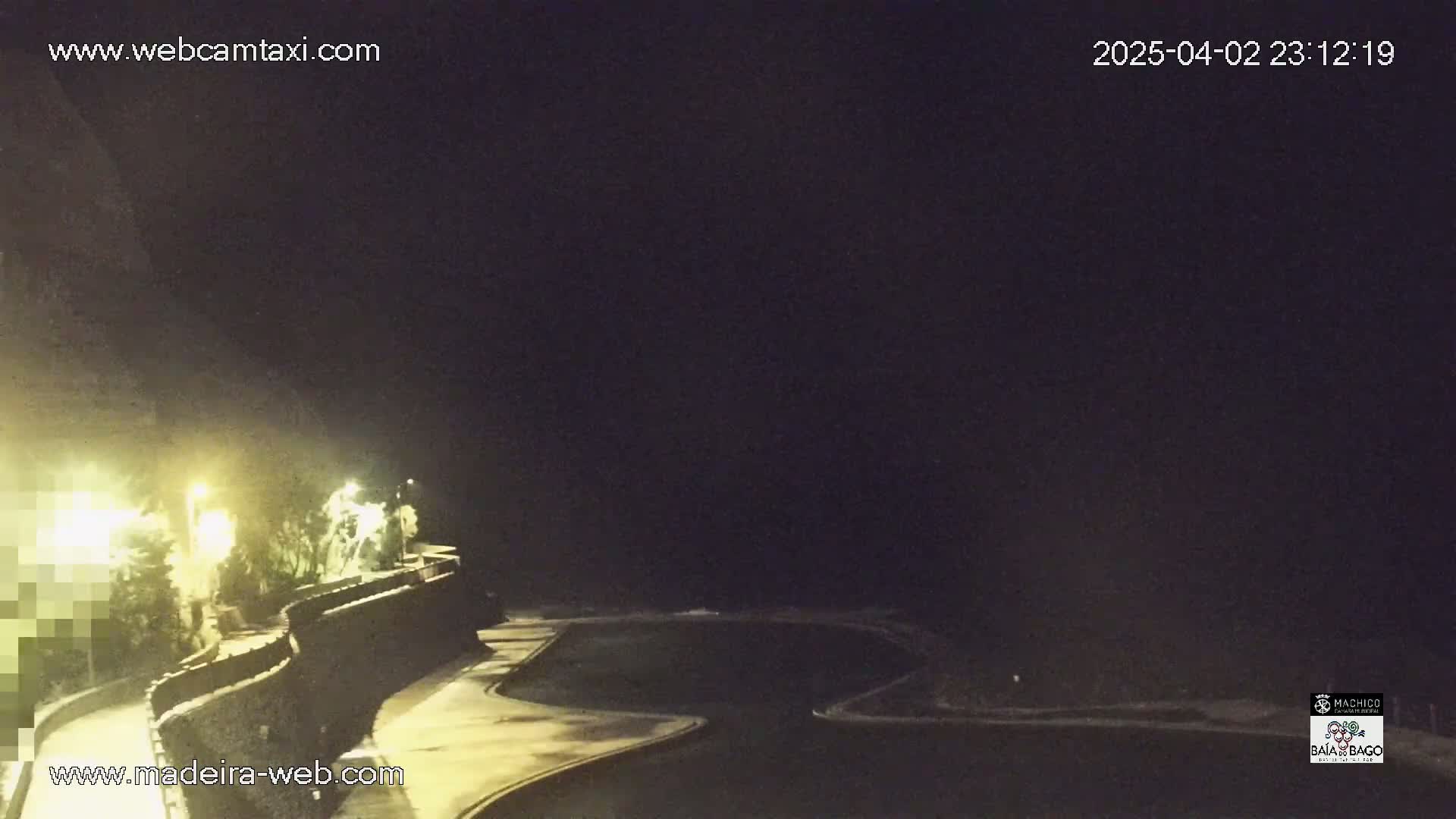 A nighttime view shows a dark ocean with a lit walkway curving along a stone wall next to it, under a clear, dark sky.