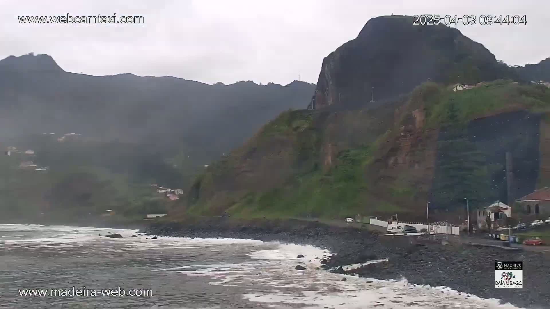A rocky coastline with dark, choppy waves under an overcast sky, showing a mountainside with some buildings along the road.
