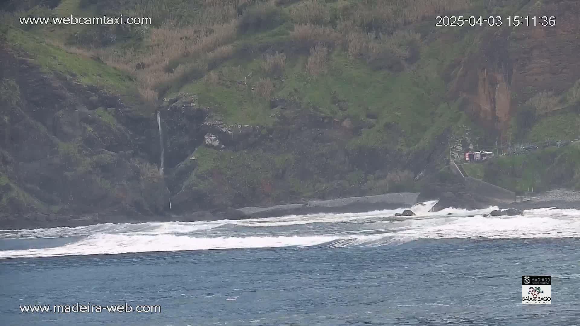 Waves crash on a rocky shore near a small waterfall cascading down a green hillside.