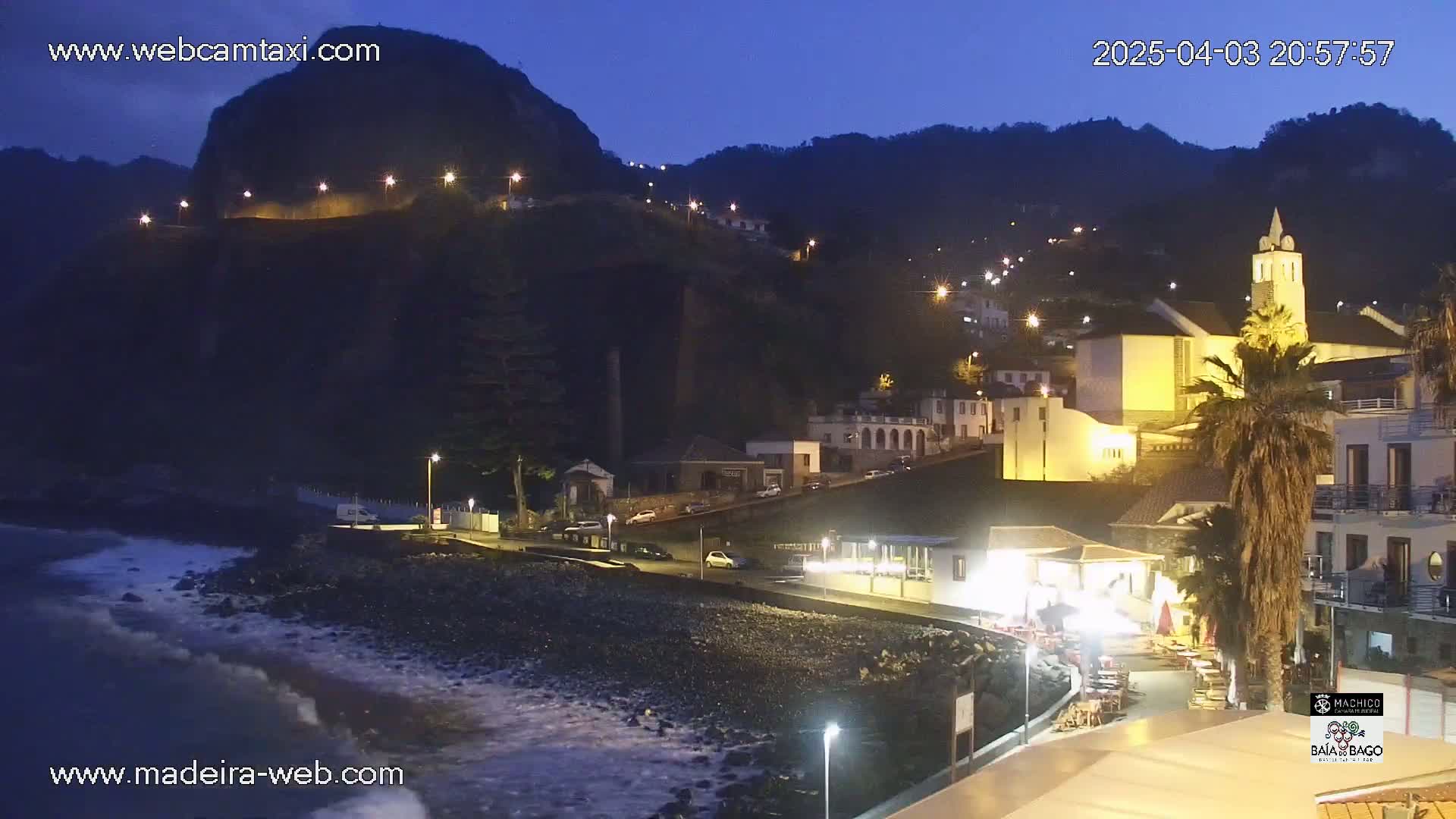A nighttime coastal town illuminated by streetlights is nestled against a dark, mountainous backdrop under a twilight sky, with gentle waves lapping at a pebble beach.