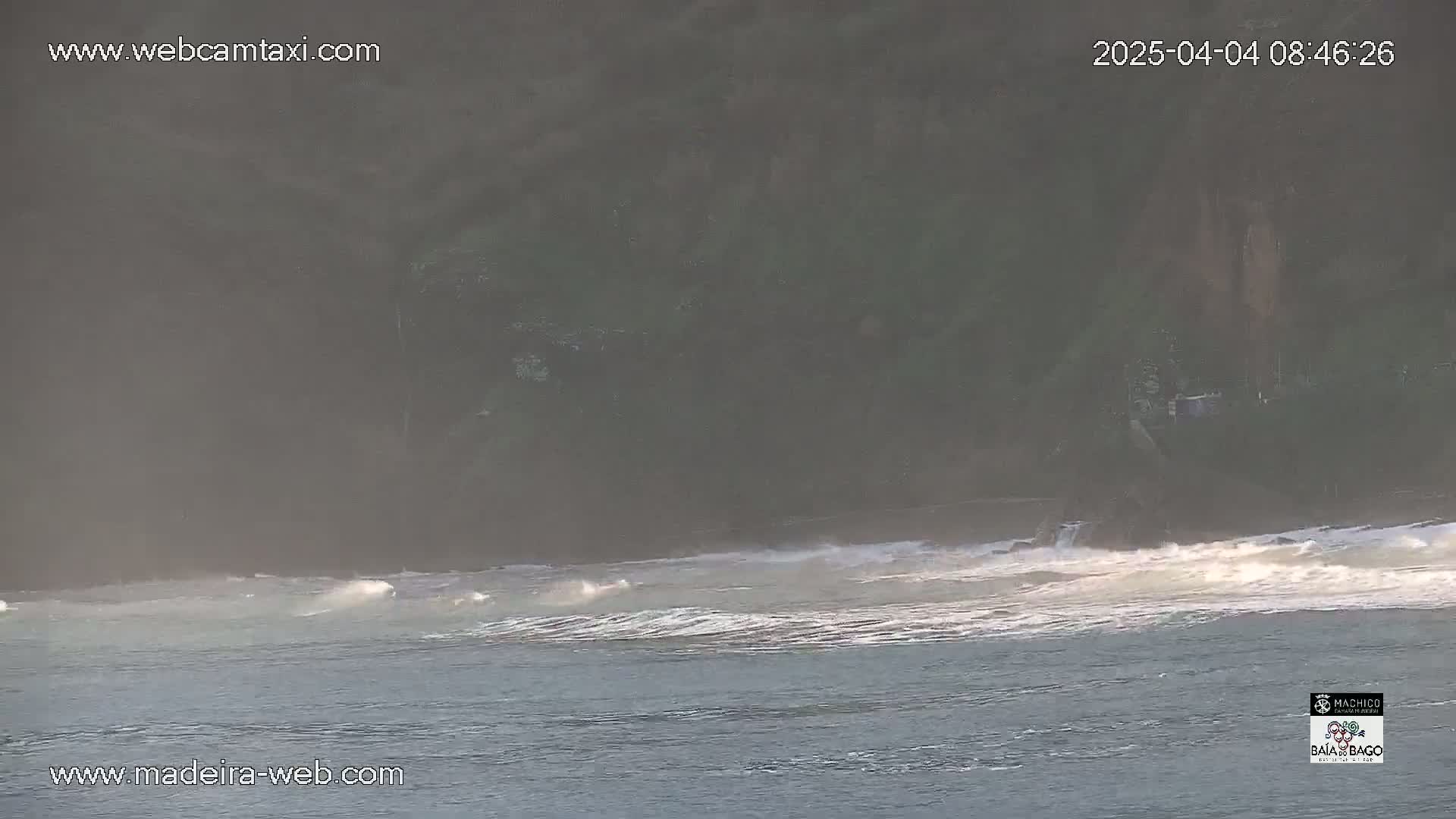 A hazy, misty view shows a dark shoreline and relatively calm ocean waves breaking gently onto a beach.