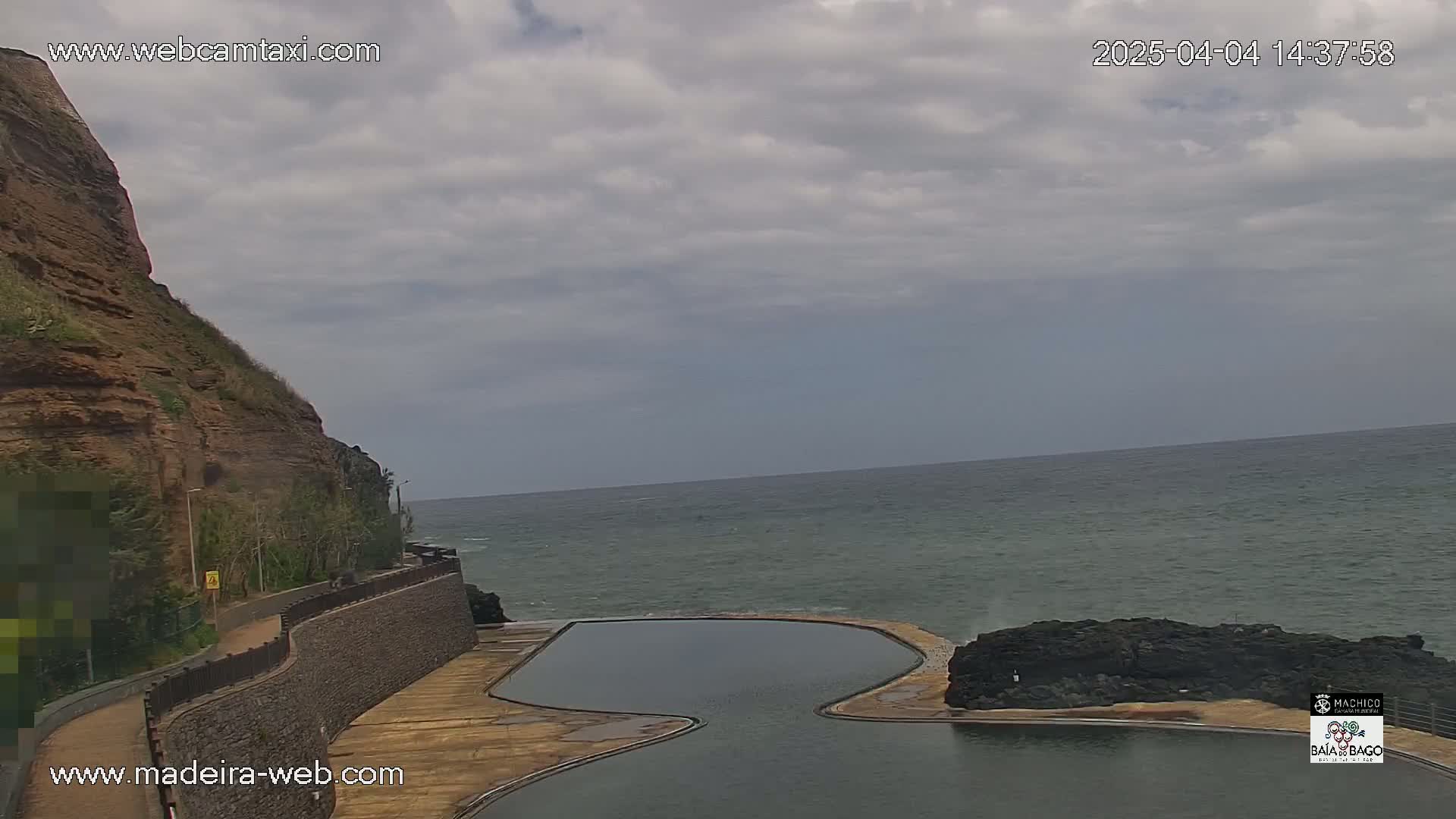 A rocky cliff overlooks a calm ocean and a kidney-shaped swimming pool under a cloudy sky.
