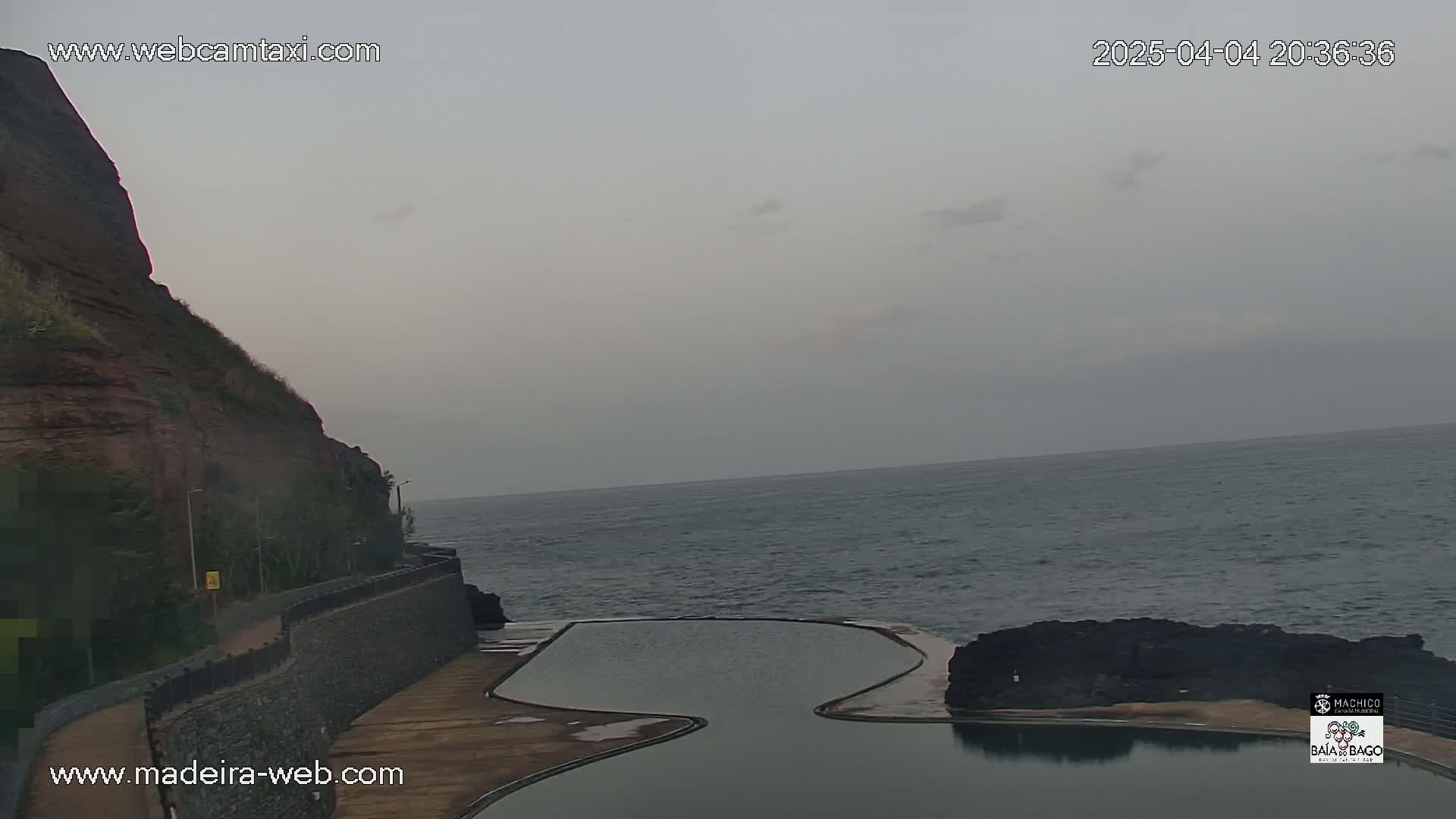A rocky cliff overlooks a calm ocean with an empty, curved swimming pool situated between the cliff and the water under an overcast sky.