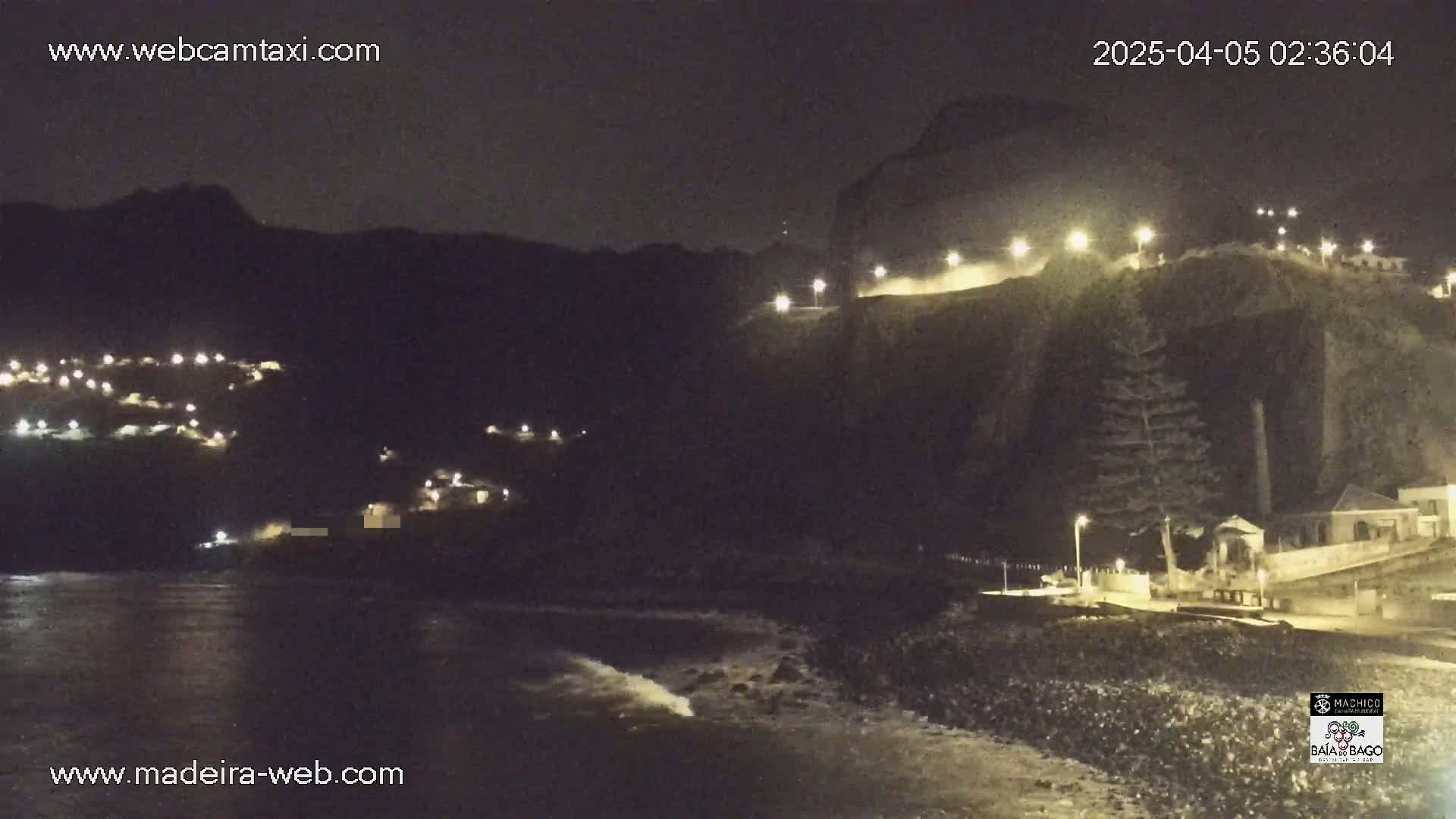 A nighttime coastal scene shows a dark, mountainous area with some lights, a rocky shoreline with gentle waves, and a few buildings illuminated along the water.