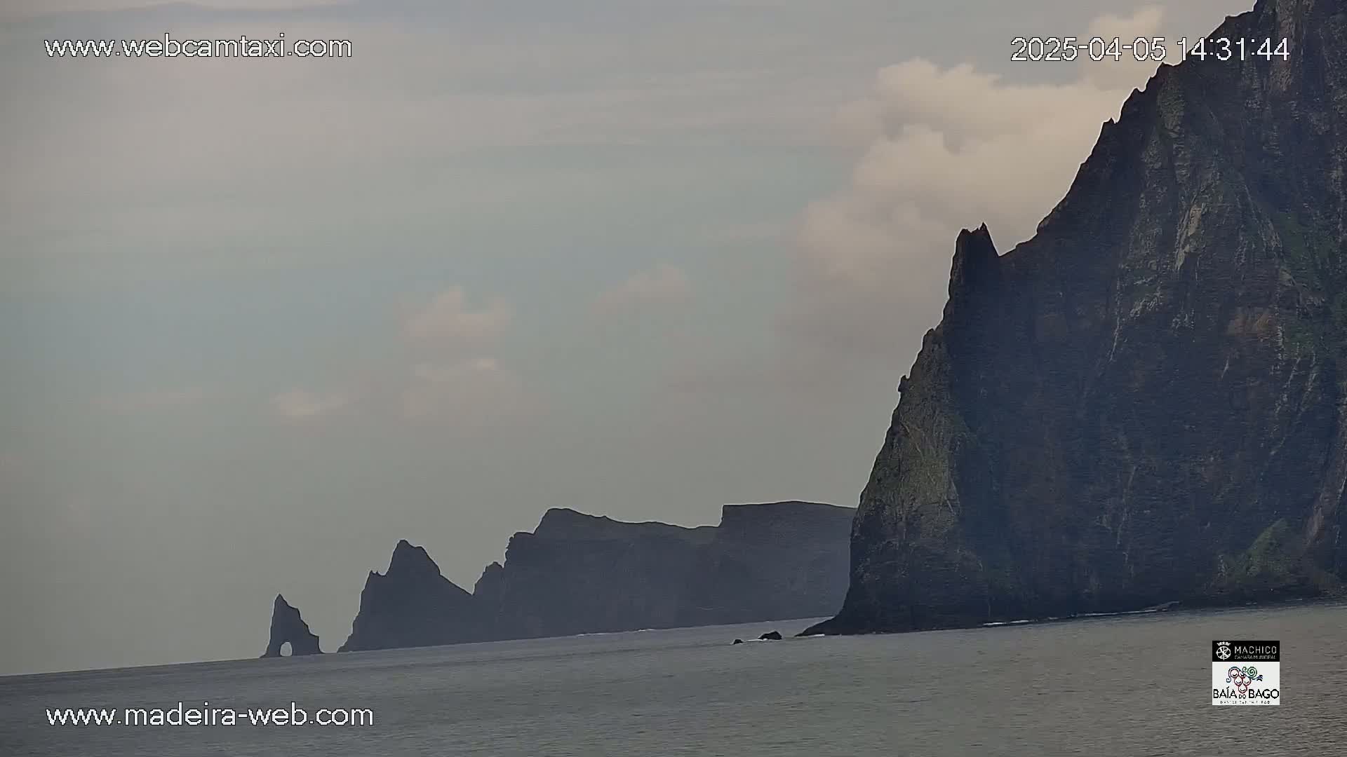 A mostly cloudy sky overlooks dark, jagged cliffs extending into a calm ocean.