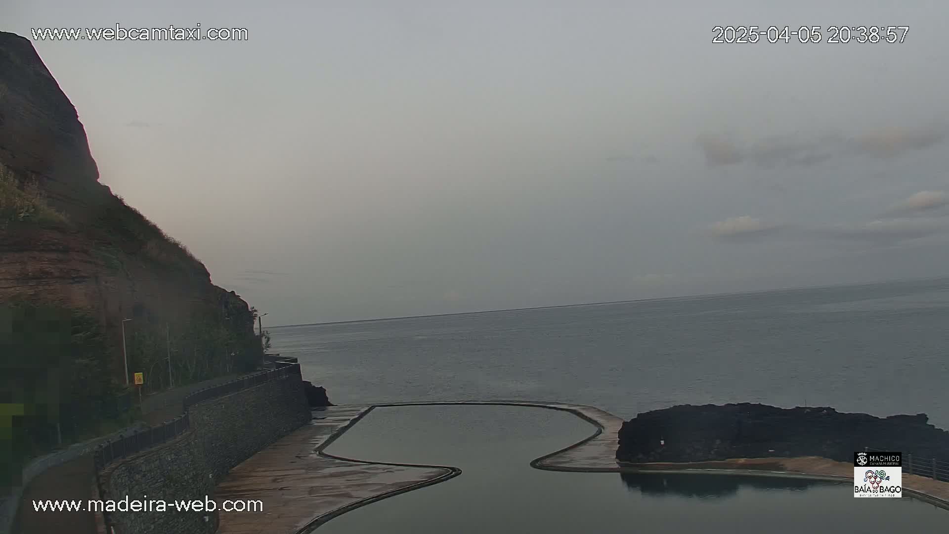 A coastal scene under an overcast sky shows a rocky cliff, a calm ocean, and a curved man-made pool near the water's edge.