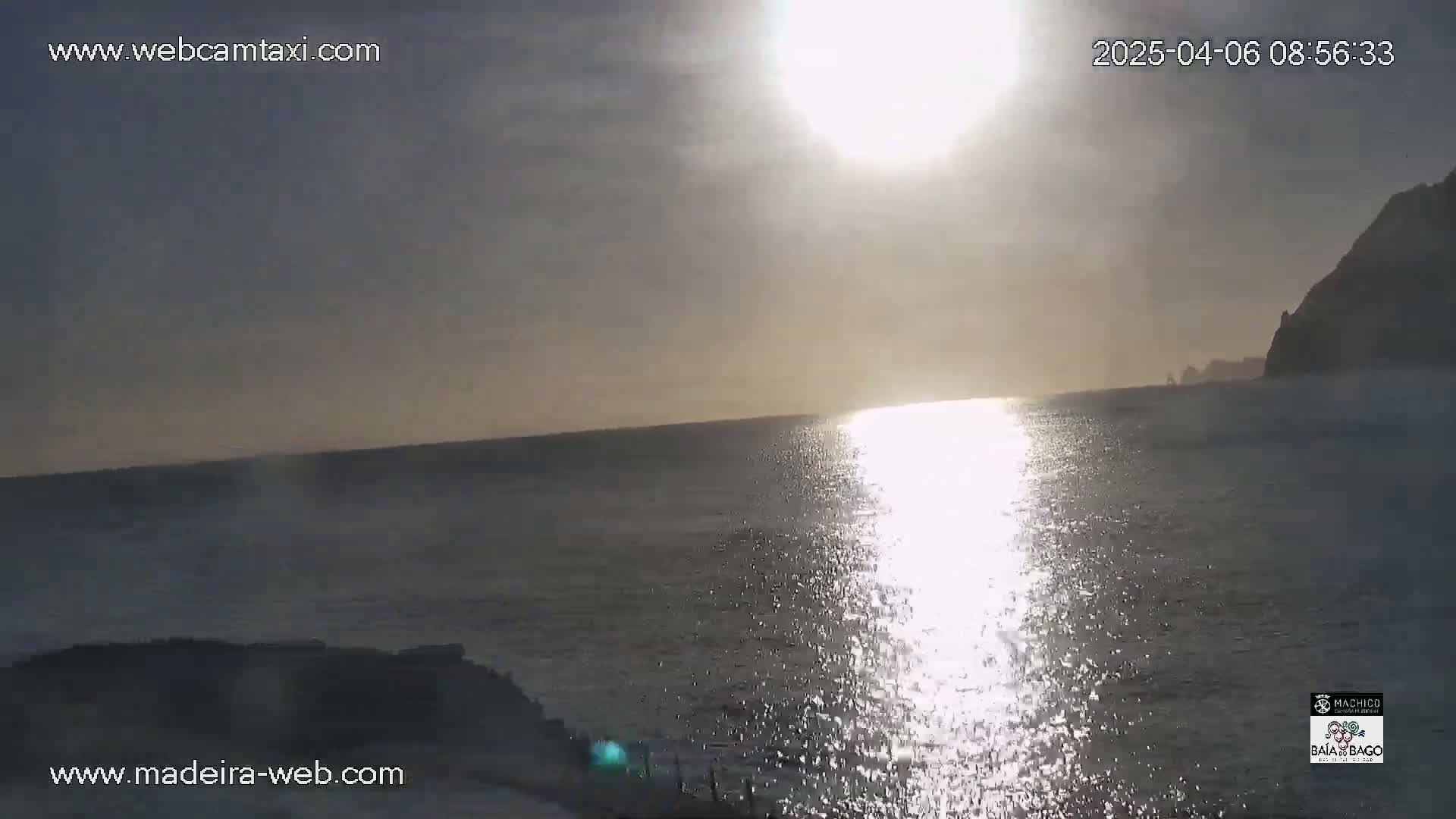 A bright sun shines over a calm ocean, with a dark cliff visible in the distance under a mostly clear sky.